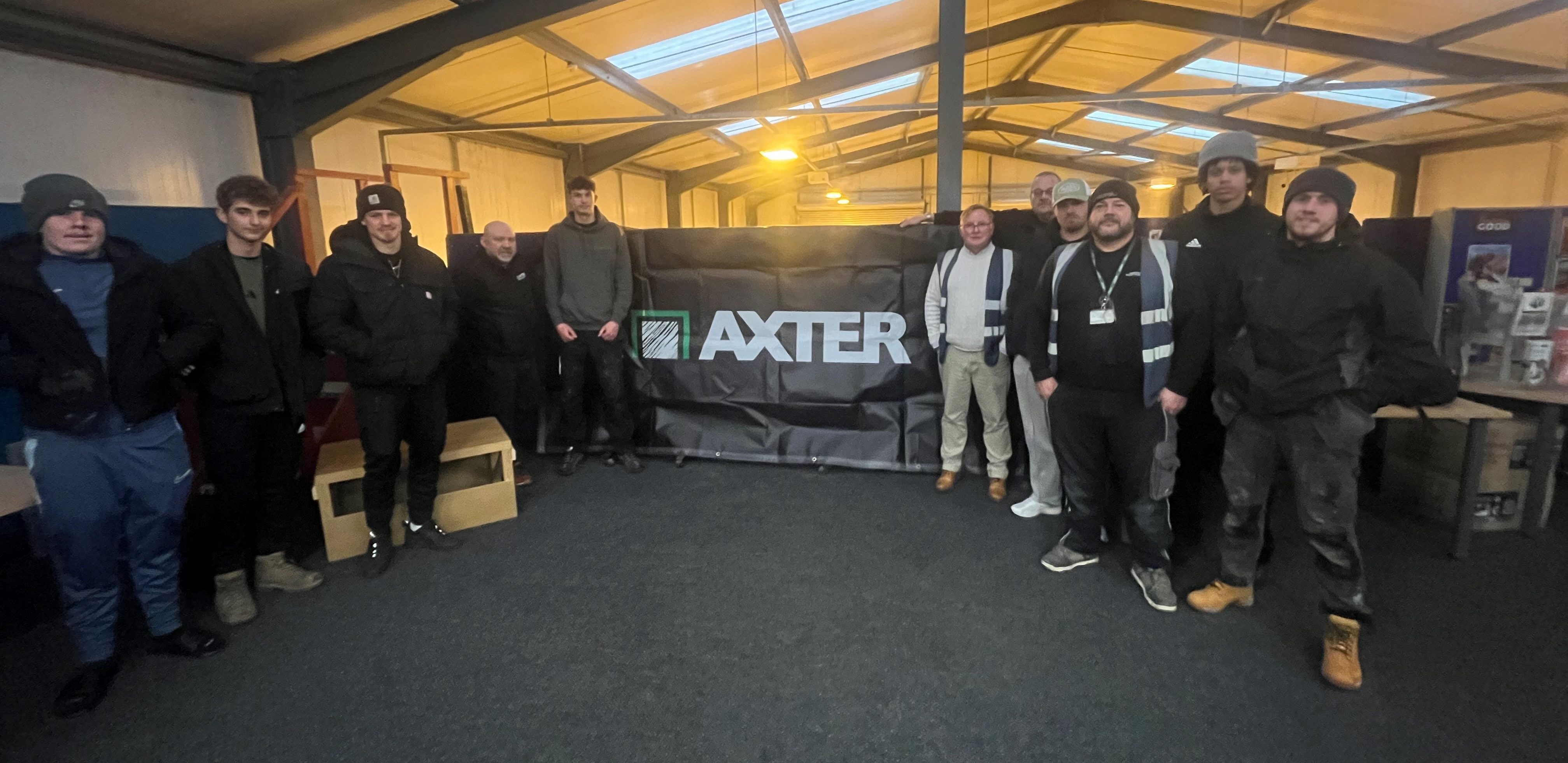 Group of people standing in warehouse with green floor, metal roof structure, and "AXTER" sign on wall behind them.