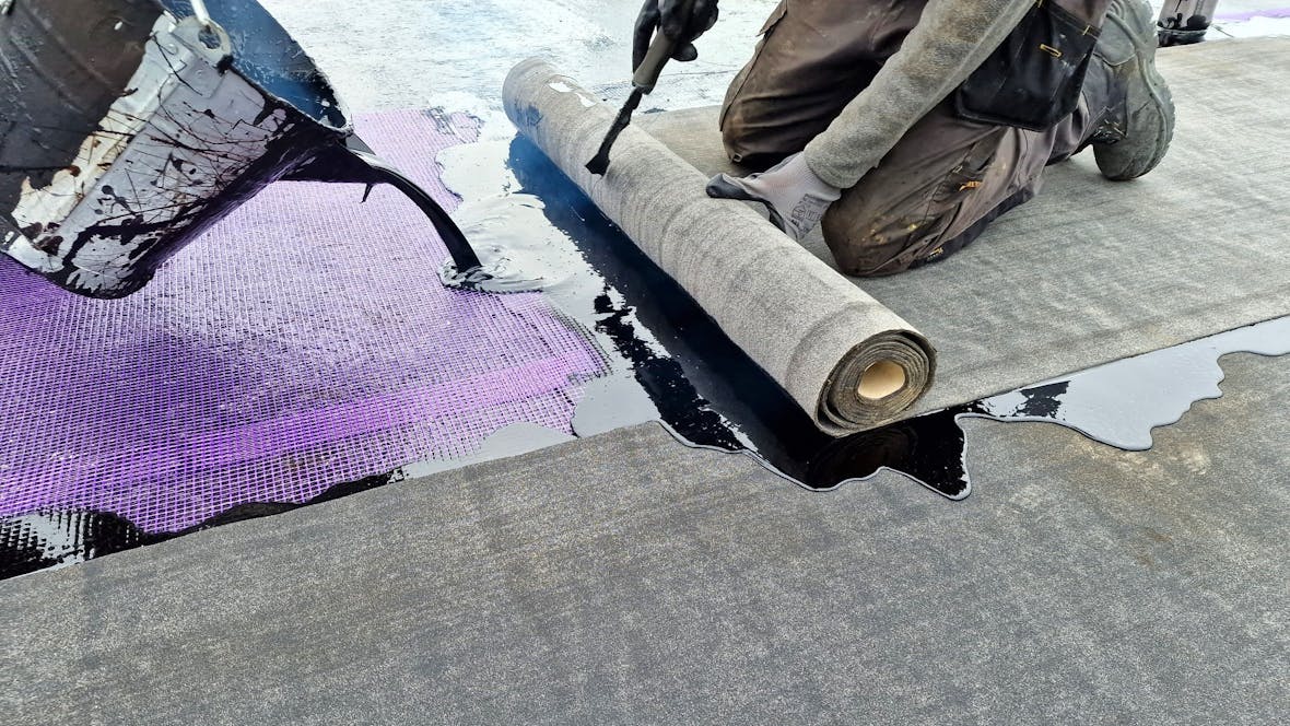 Person kneeling on concrete surface applying black adhesive or sealant from a roll to purple mesh material.