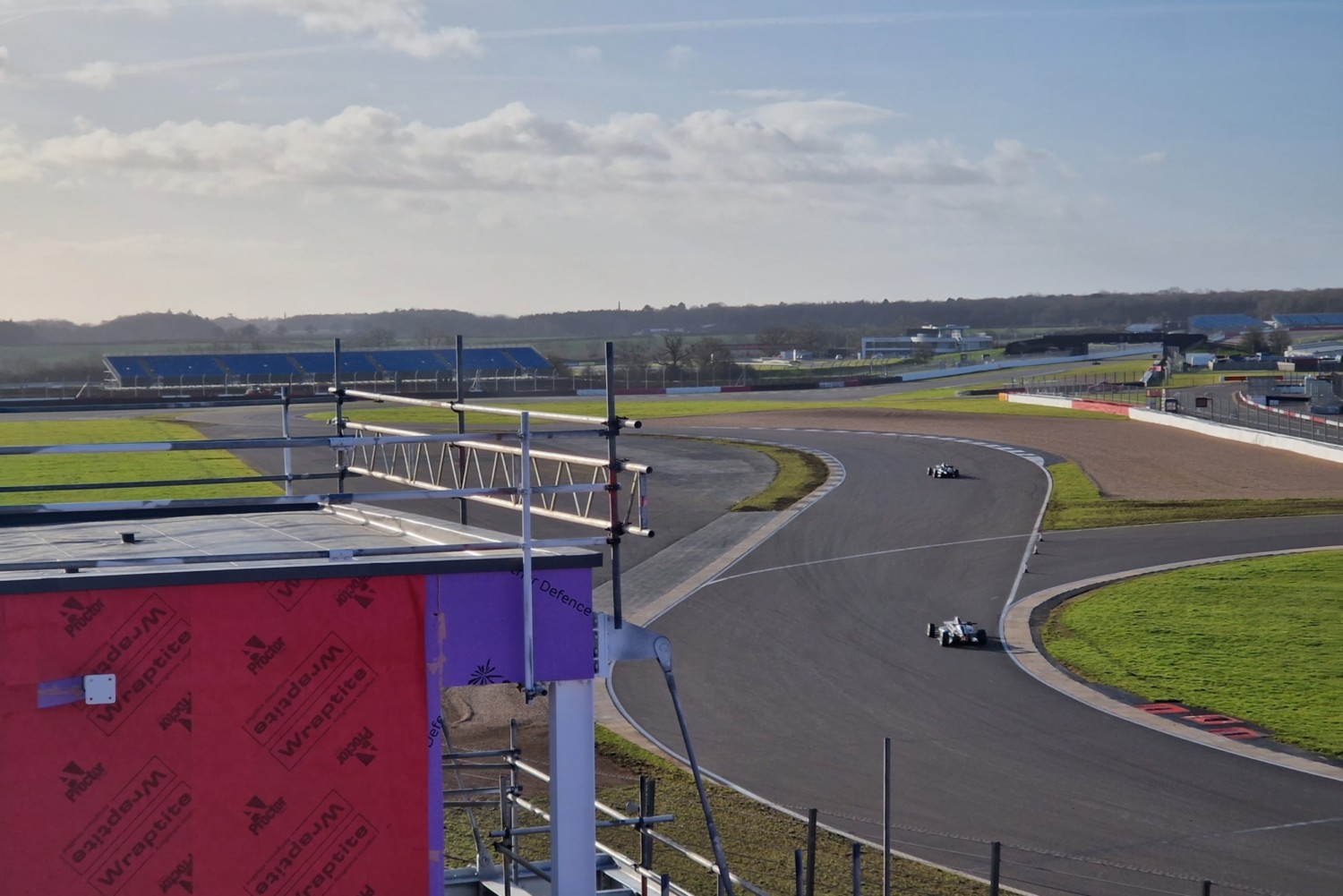 Race circuit with curved tarmac track, green grass verges, red and purple grandstand structure, and cloudy sky overhead.