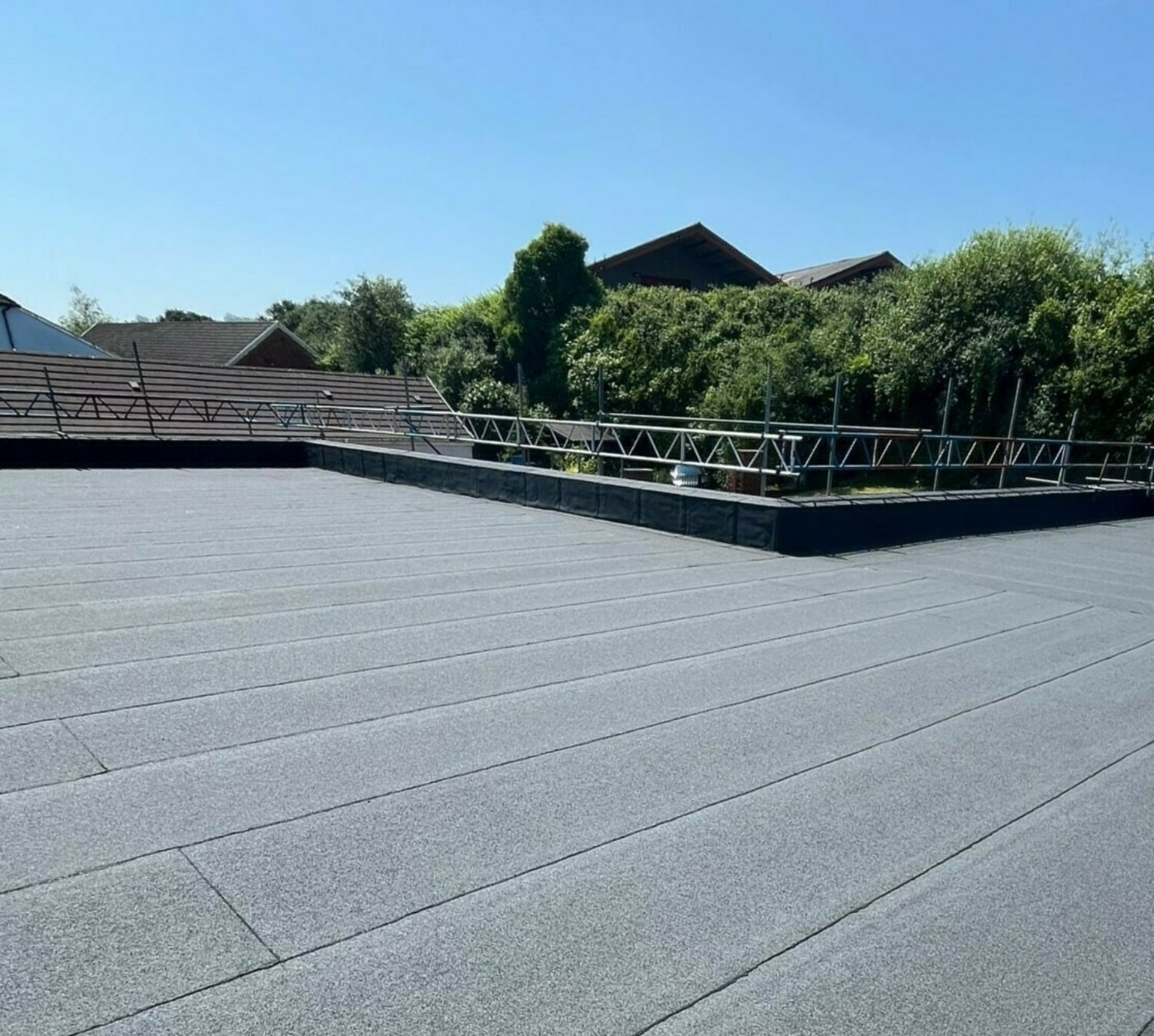 Grey flat roof with white metal safety railings, residential houses and green trees visible in background under blue sky.