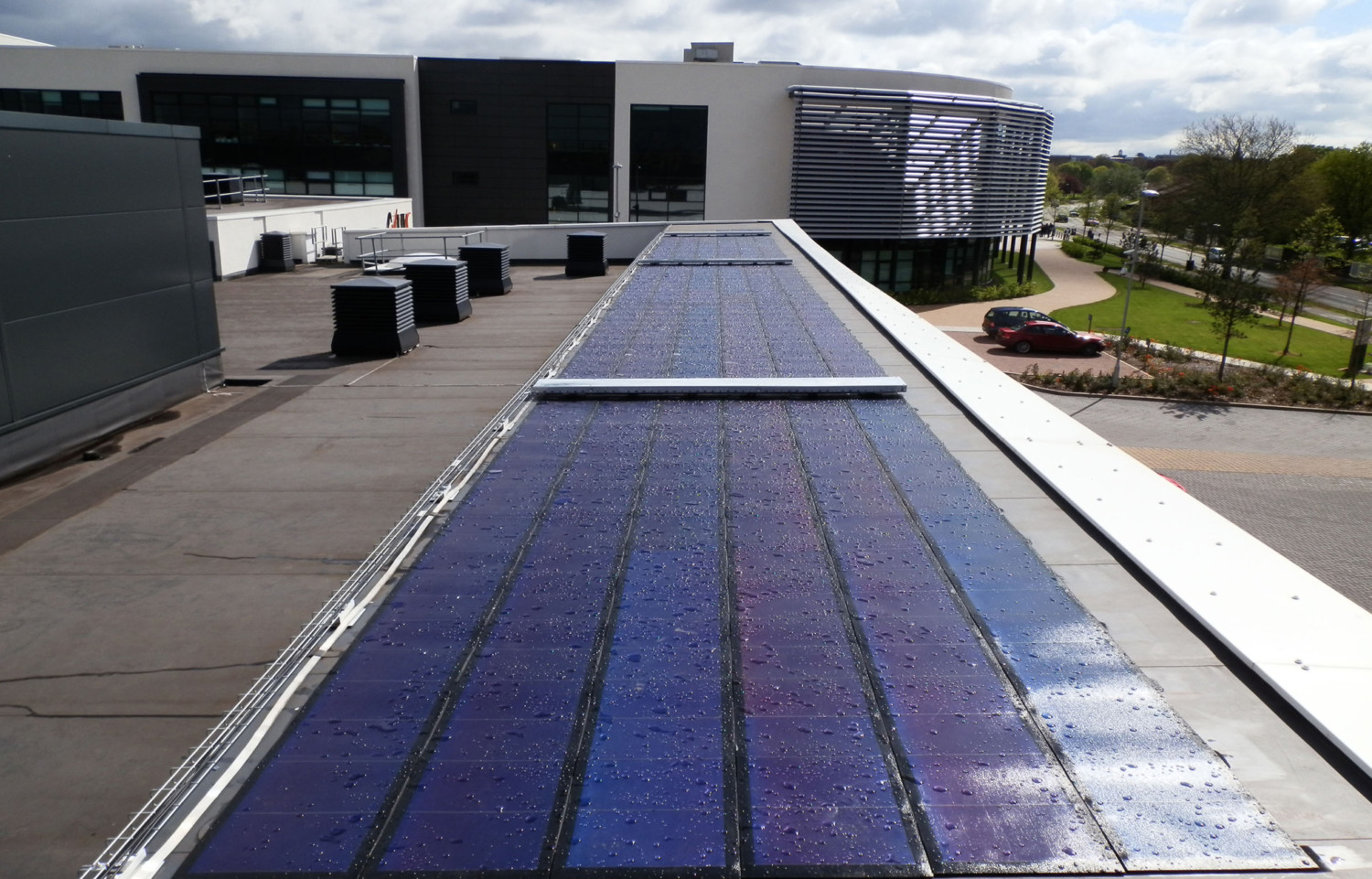 Blue solar panels installed on flat rooftop of modern grey building with glass windows and metal cladding under cloudy sky.