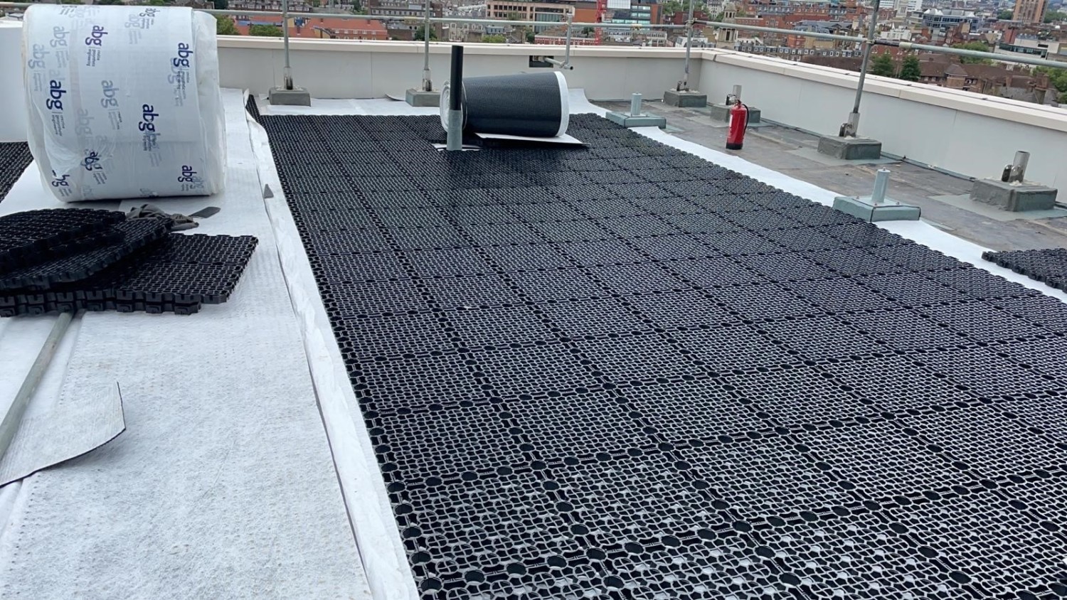 Black honeycomb drainage matting covers most of flat roof surface, with white insulation sections and cylindrical equipment visible.