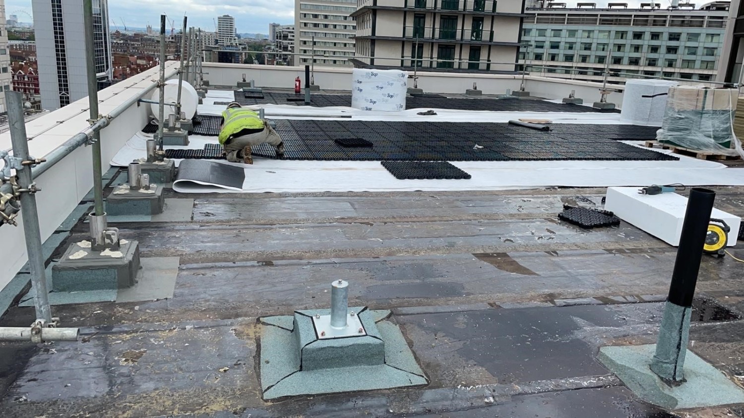 Rooftop construction site with grey concrete surface, white panels, green equipment, and city buildings in background.