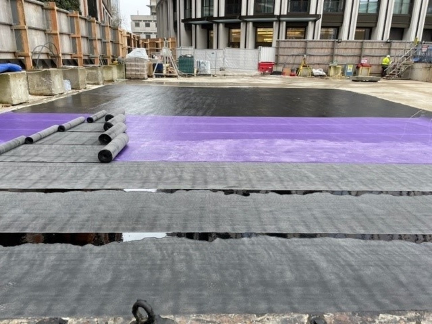 Building site with purple and green waterproofing membrane laid on concrete surface, construction materials scattered around.