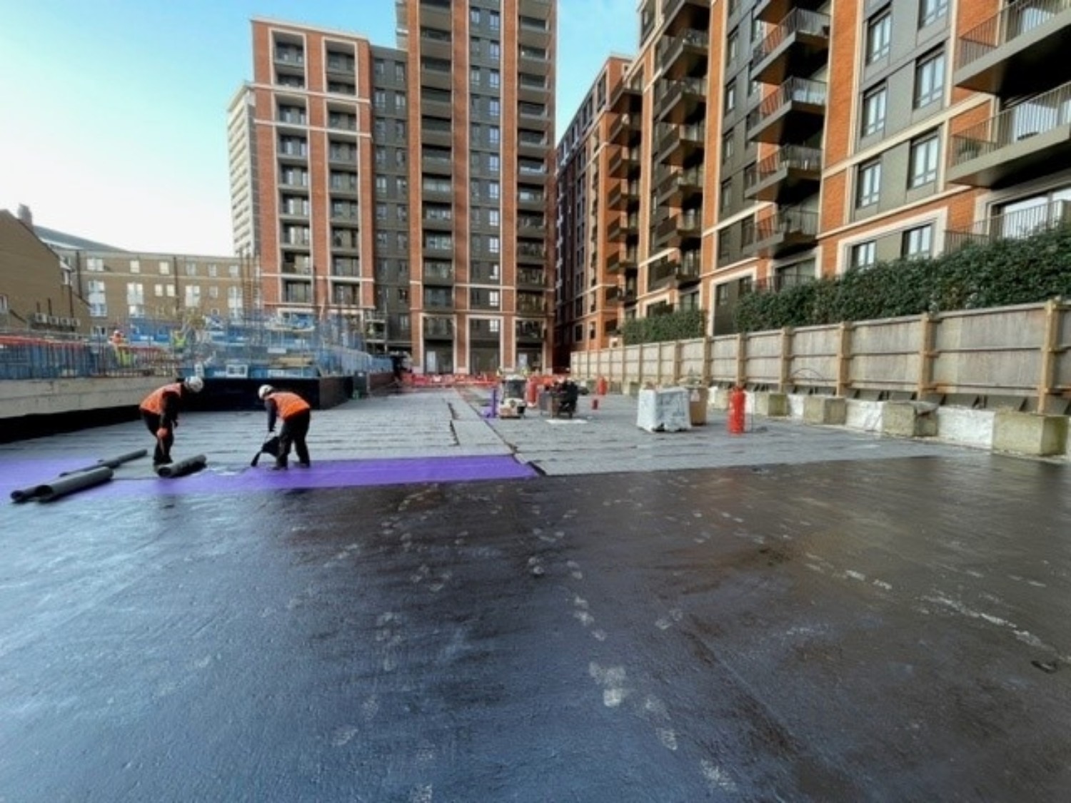 Two workers in high-vis jackets laying purple material on dark surface between modern brick apartment blocks under clear sky.