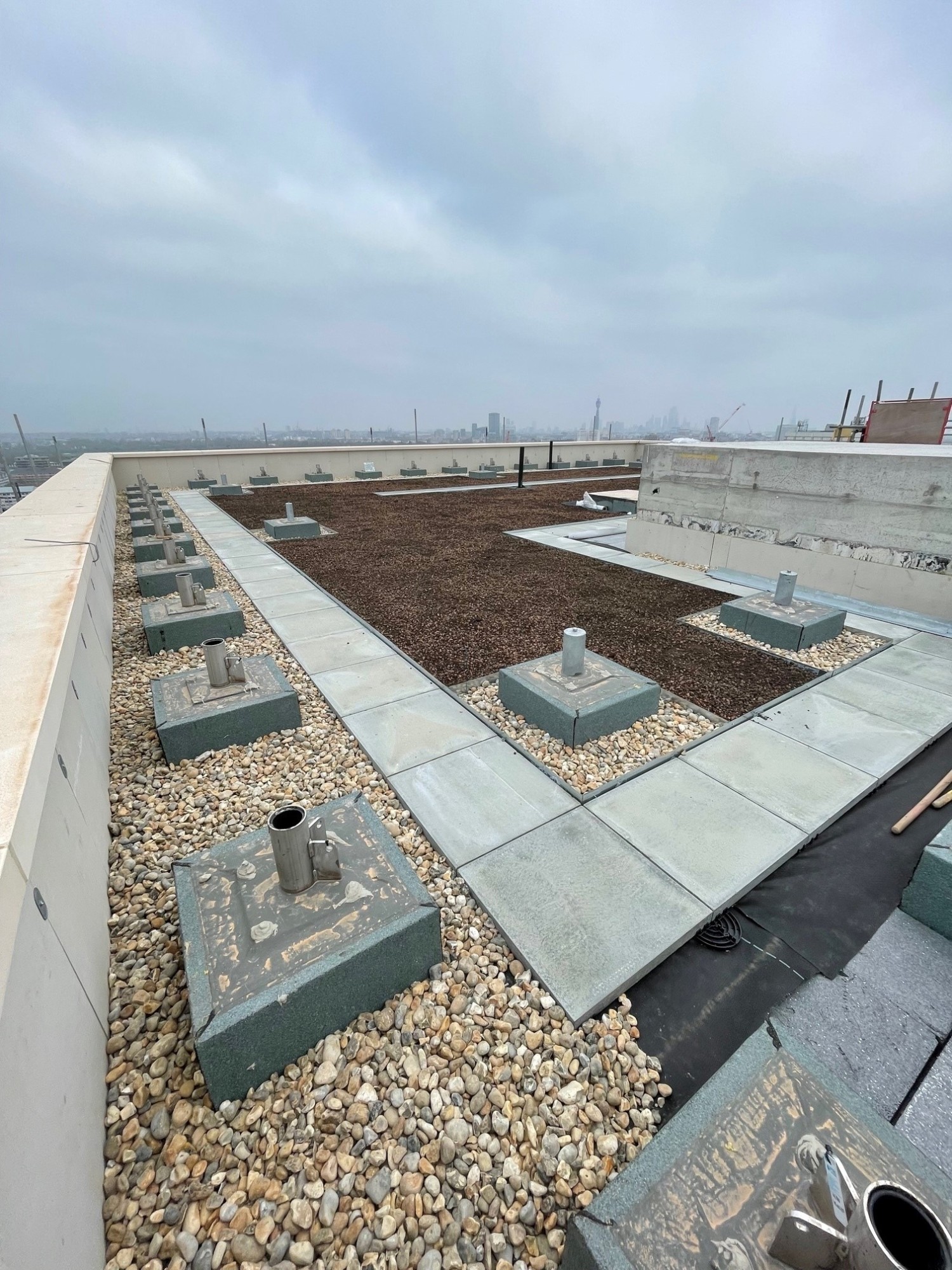 Rooftop with brown soil sections, grey concrete walkways, scattered pebbles, and mechanical equipment under cloudy sky.