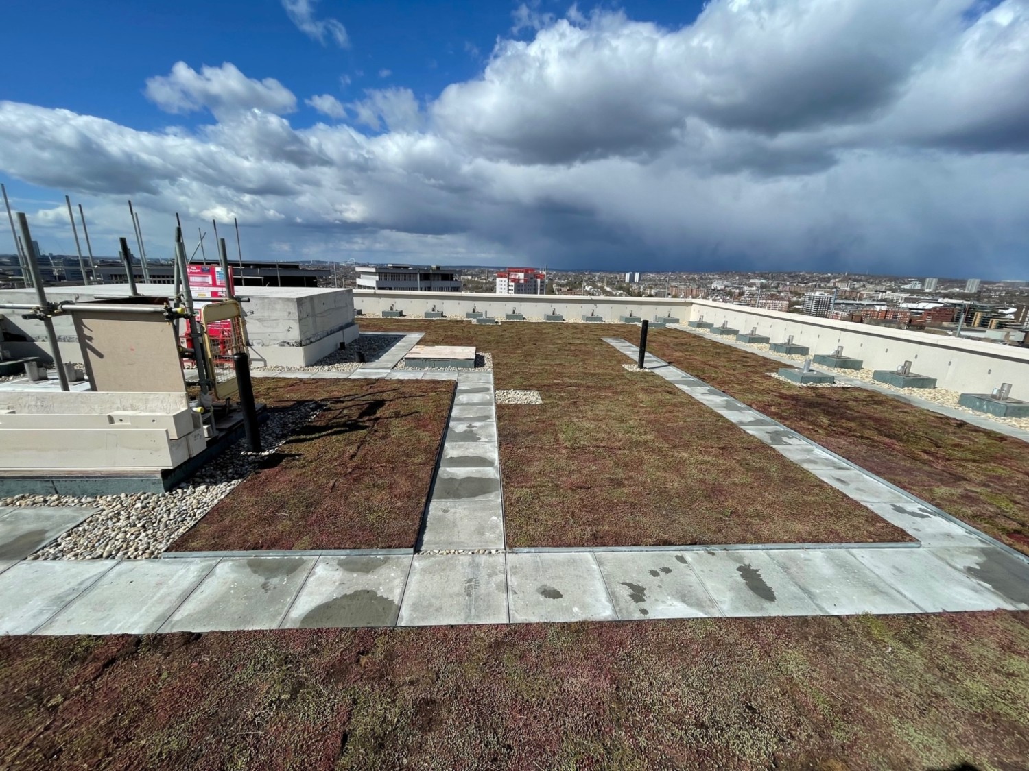 Rooftop construction site with brown soil patches, grey concrete sections, and construction equipment under blue sky with white clouds.