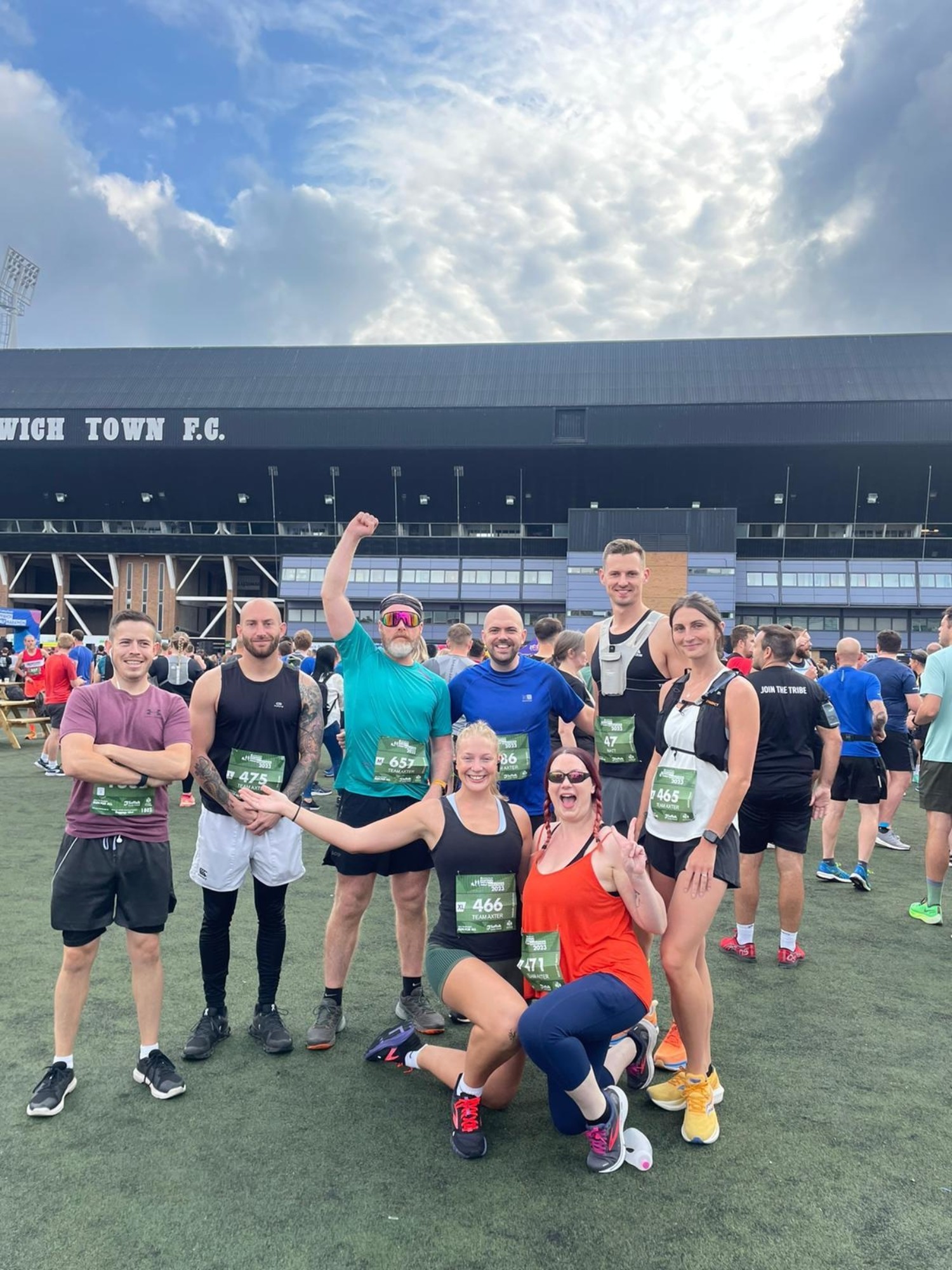 Group of runners in athletic wear posing with race medals outside football stadium under cloudy sky.