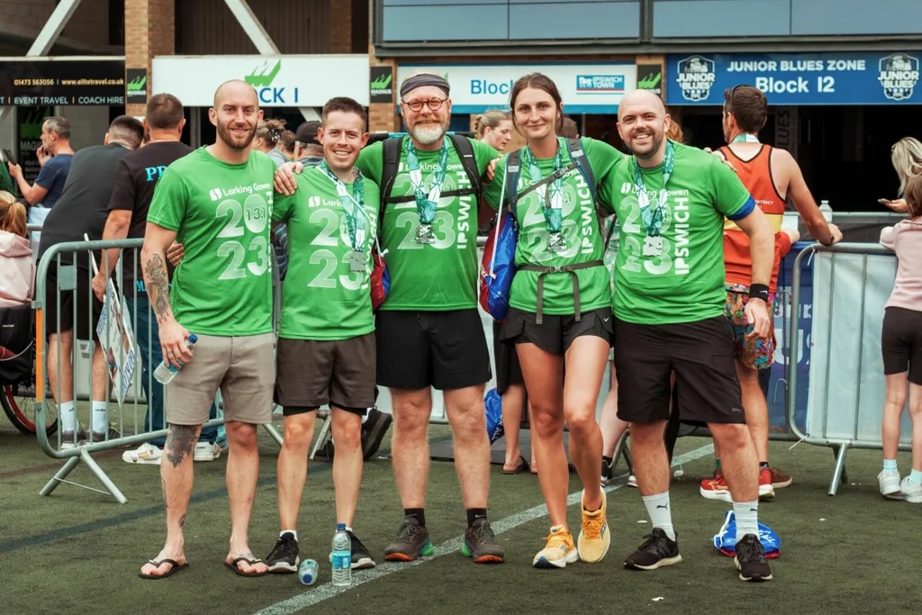 Six people in matching green t-shirts pose together at outdoor event, metal barriers and crowd visible in background.