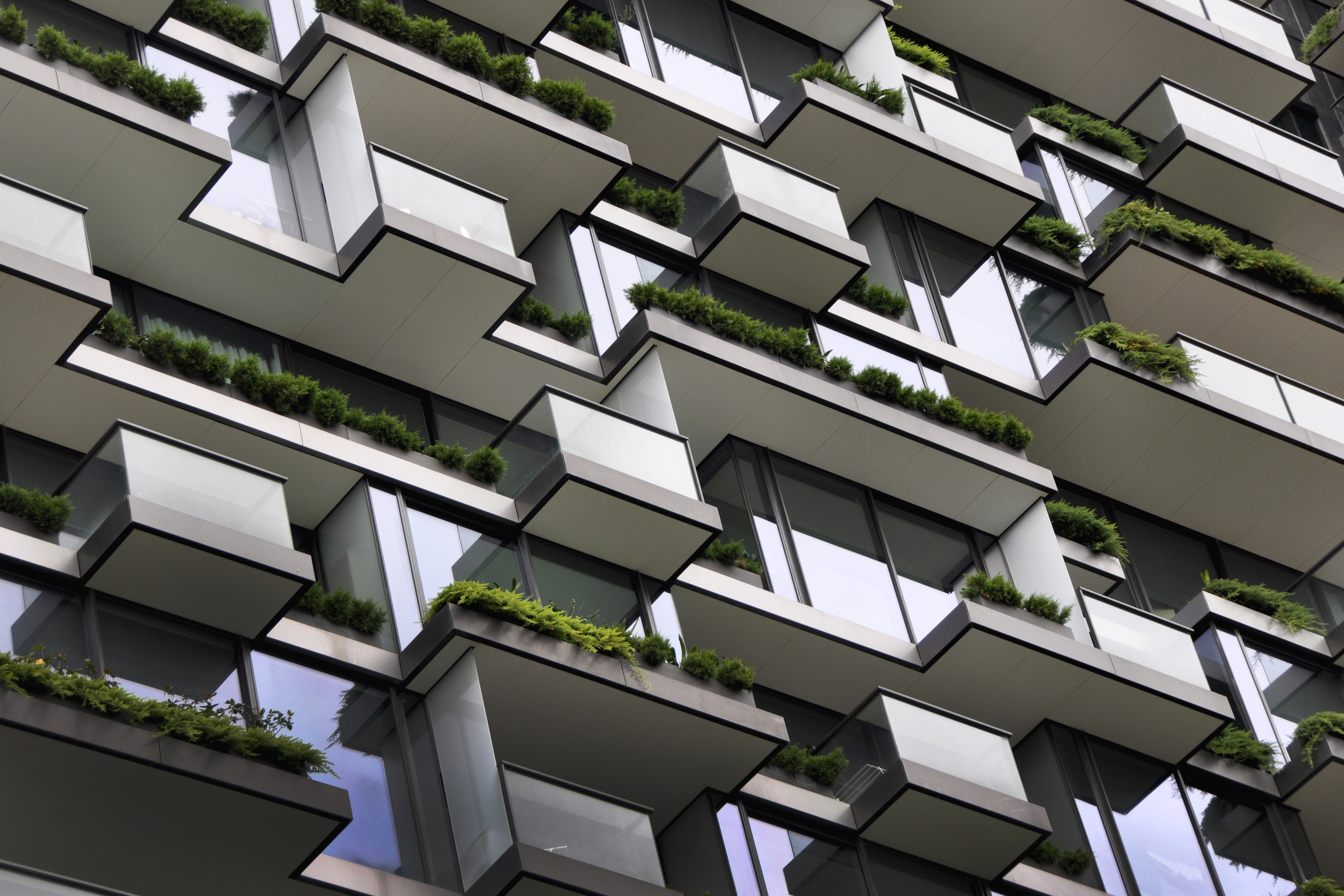 Modern building facade with angular white balconies, grey concrete structure, and green plants on terraces in geometric pattern.