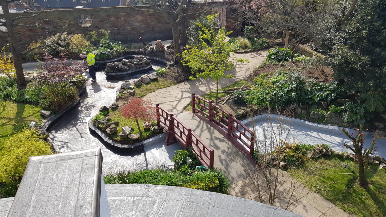 Japanese garden with curved wooden bridge over pond, stone pathways, varied green foliage, and water features viewed from above.