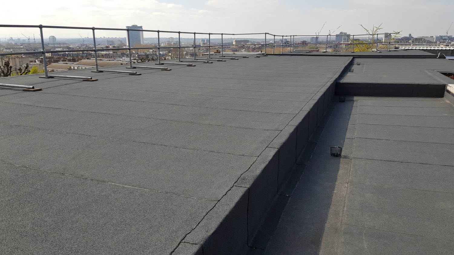 Flat rooftop with grey membrane surface, metal railings along edge, and dark rectangular opening. Industrial buildings visible in background.