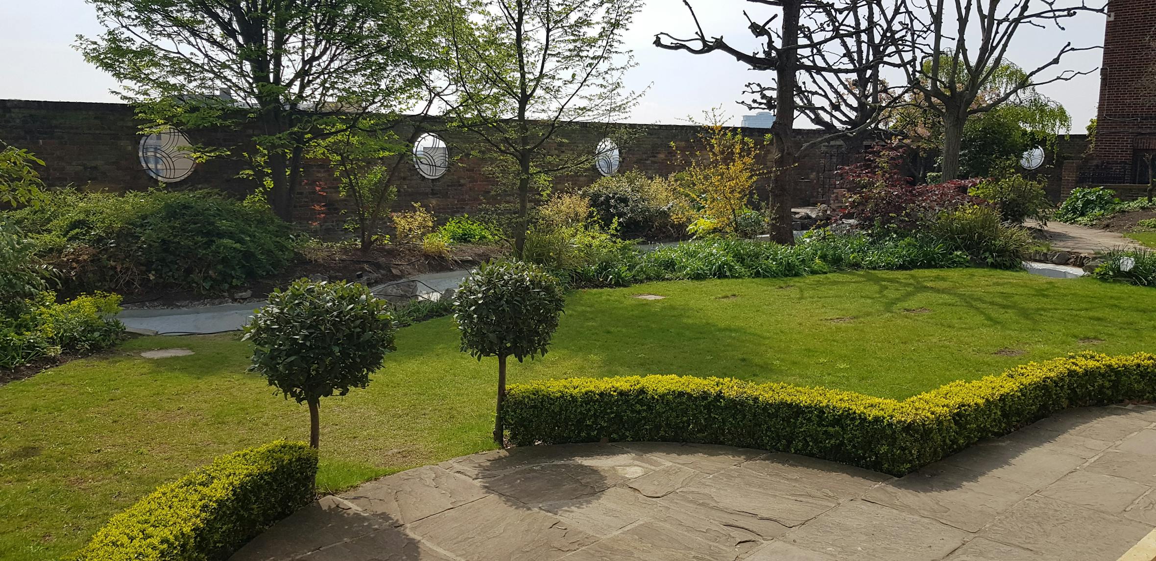 Formal garden with geometric green lawn sections bordered by low hedges, two small topiary trees, and mature trees behind stone walls.