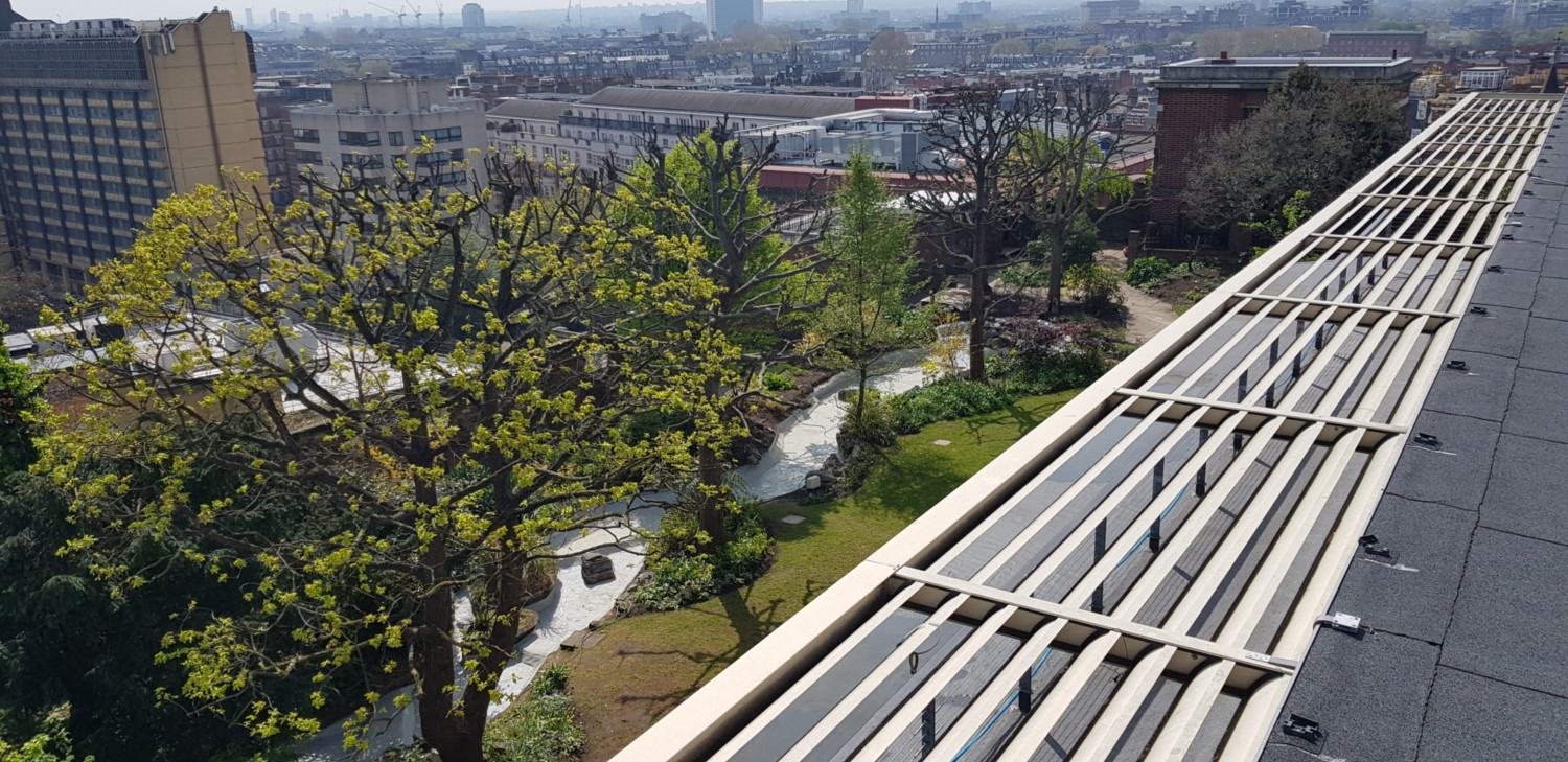 Aerial view of urban cityscape with green trees, buildings, and white striped rooftop structure in foreground extending diagonally.