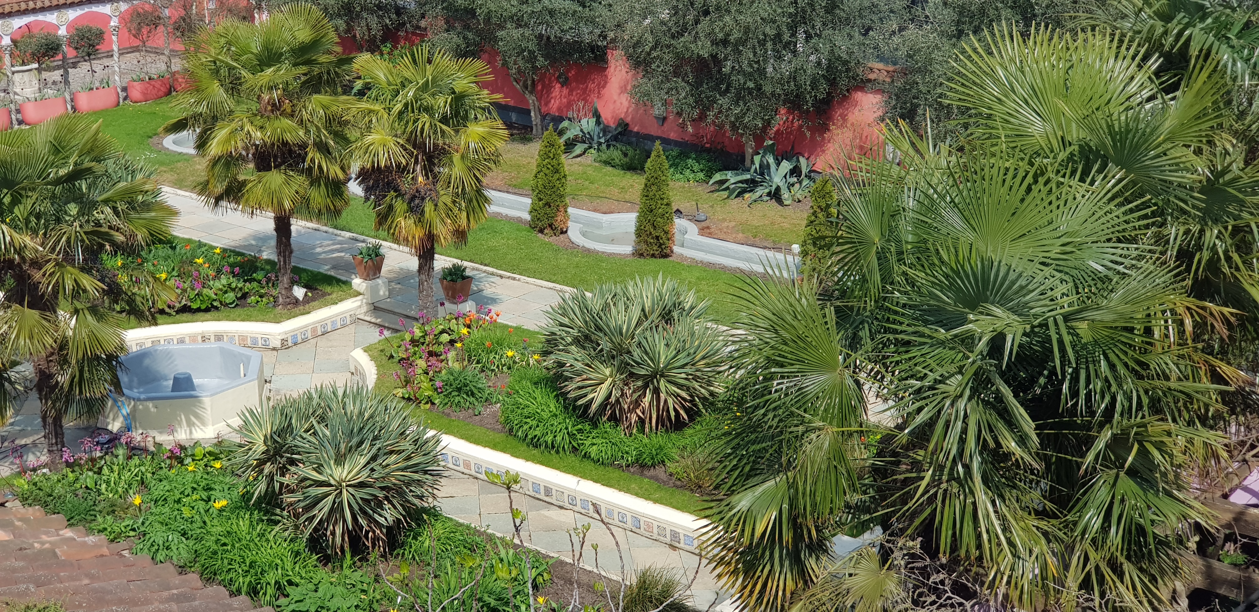 Aerial view of tropical garden with palm trees, curved pathways, green lawns, and terracotta-coloured buildings in background.