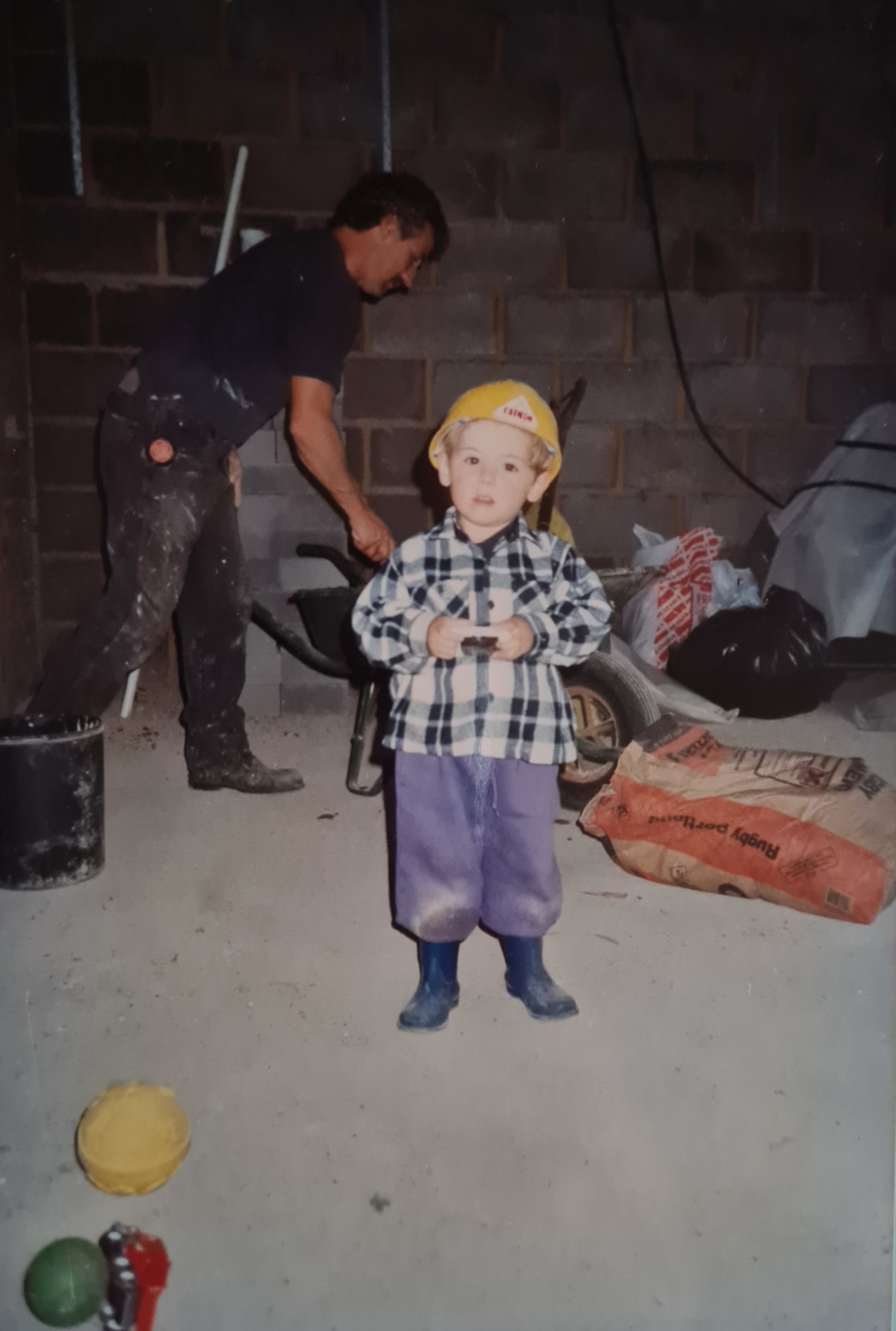 Child in yellow hard hat and chequered shirt standing in garage whilst man works on motorcycle in background