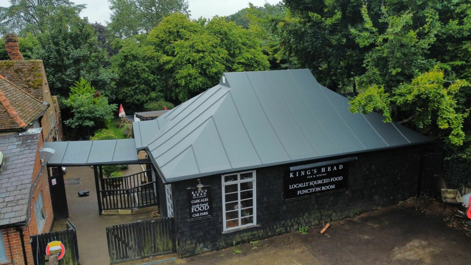 Dark grey building with pyramid-shaped metal roof surrounded by green trees, with traditional brick houses visible nearby.