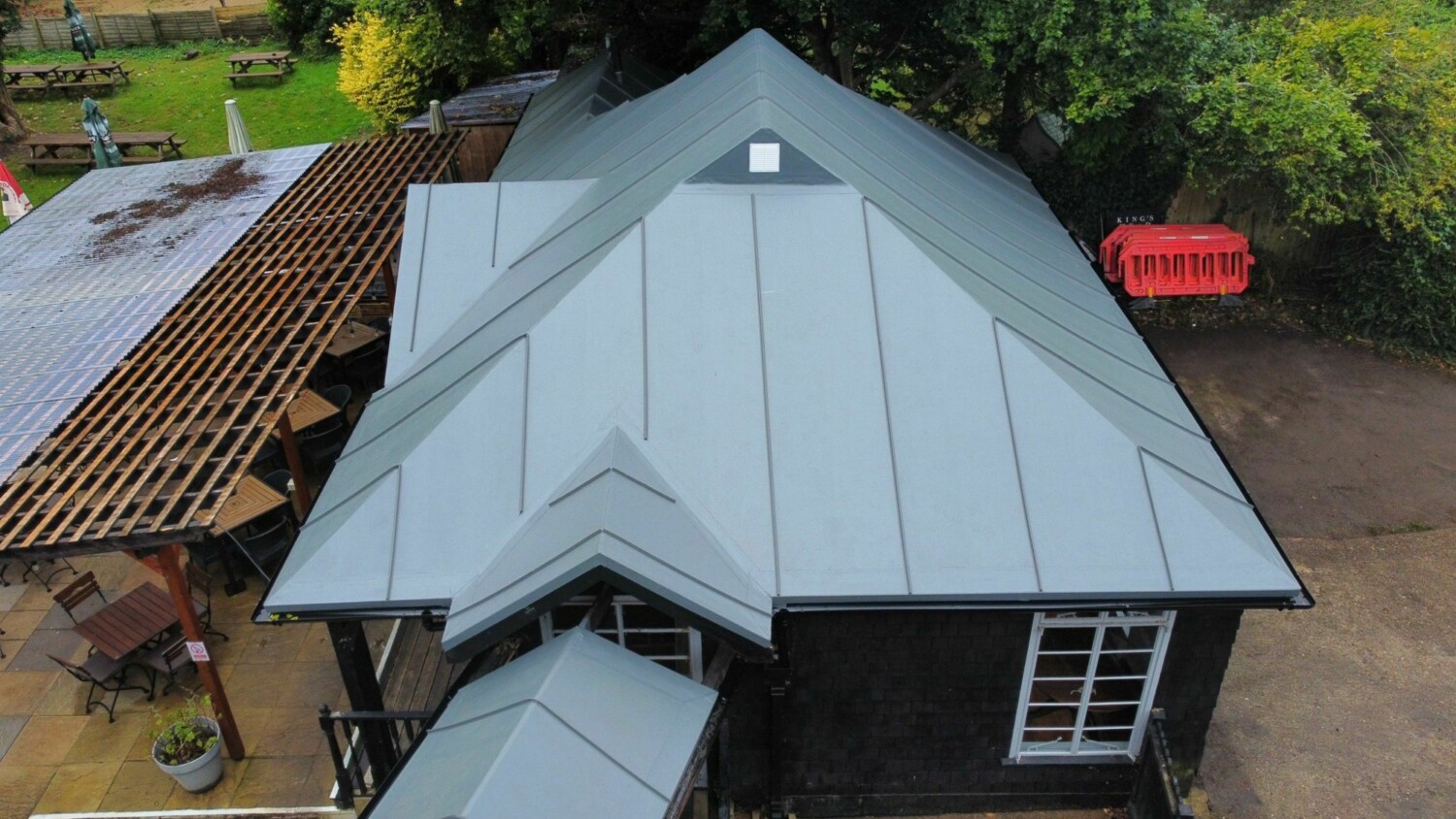 Aerial view of house with grey metal roof, brown tiled roof section, white walls, surrounded by green trees and lawn.