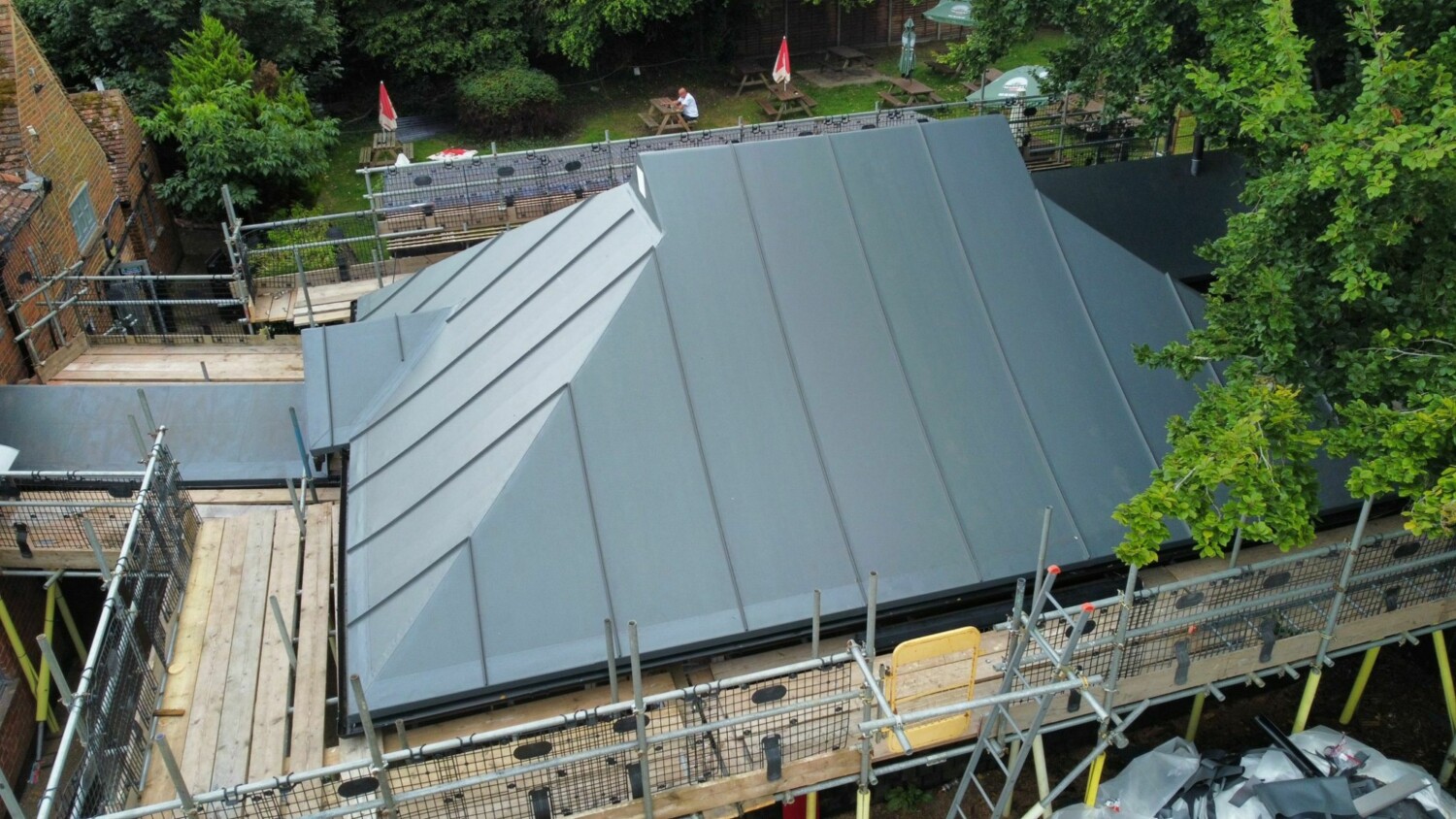 Aerial view of grey metal roof with scaffolding around building under construction, surrounded by green trees and neighbouring properties.