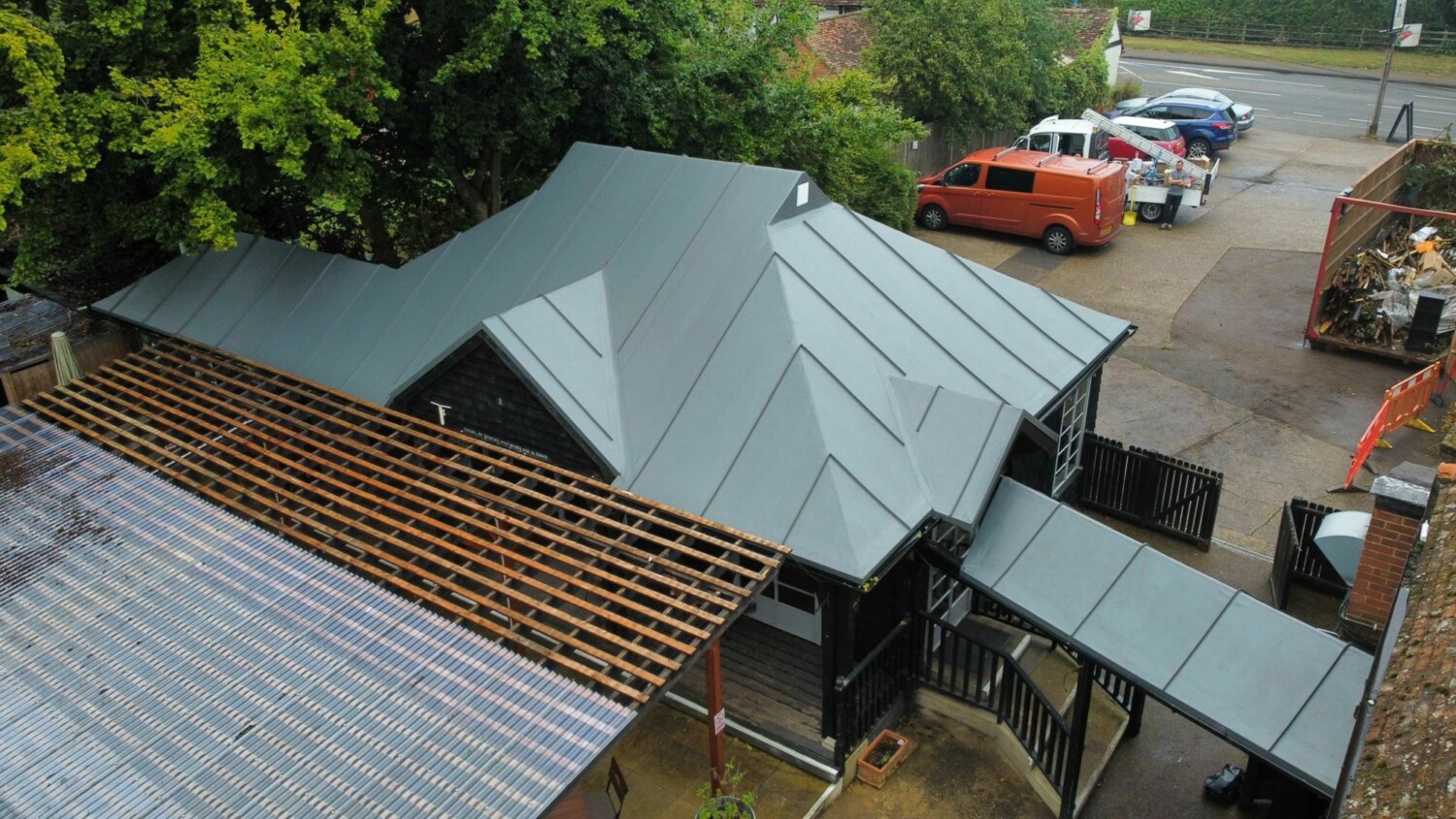 Aerial view of building with green metal roofing, exposed wooden rafters, and construction vehicles in car park surrounded by trees.