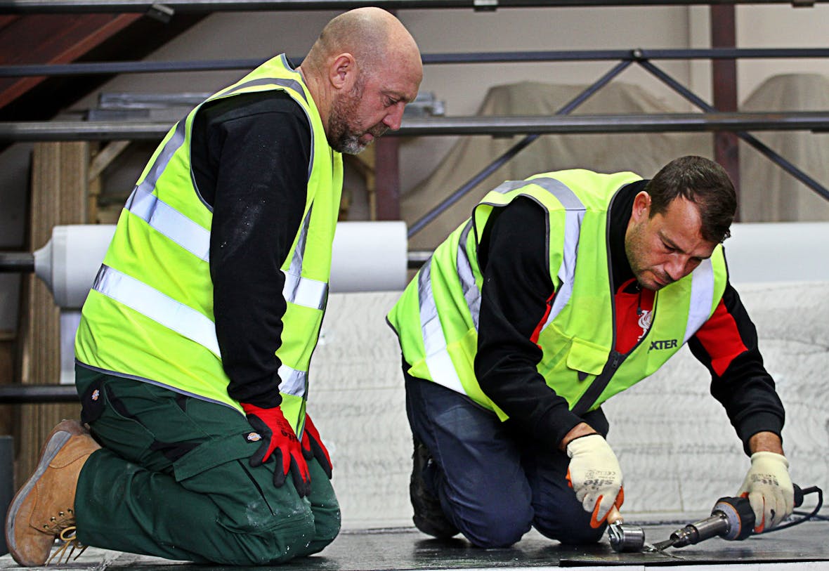Two construction workers in high-vis yellow jackets kneeling on concrete floor, working with tools near metal scaffolding.