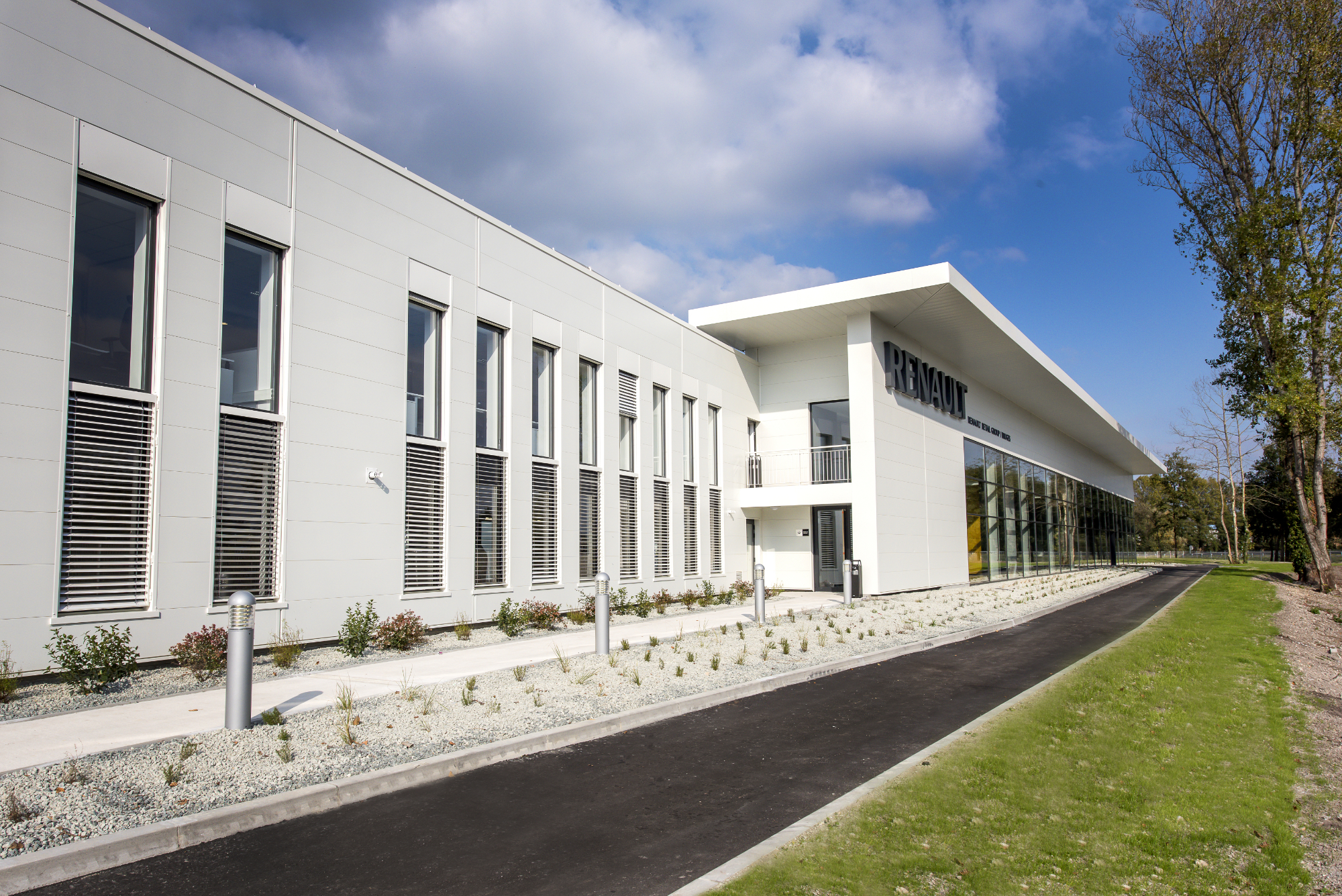 Modern white two-storey building with horizontal windows, metal shutters, and glass entrance section under blue cloudy sky with lawn.
