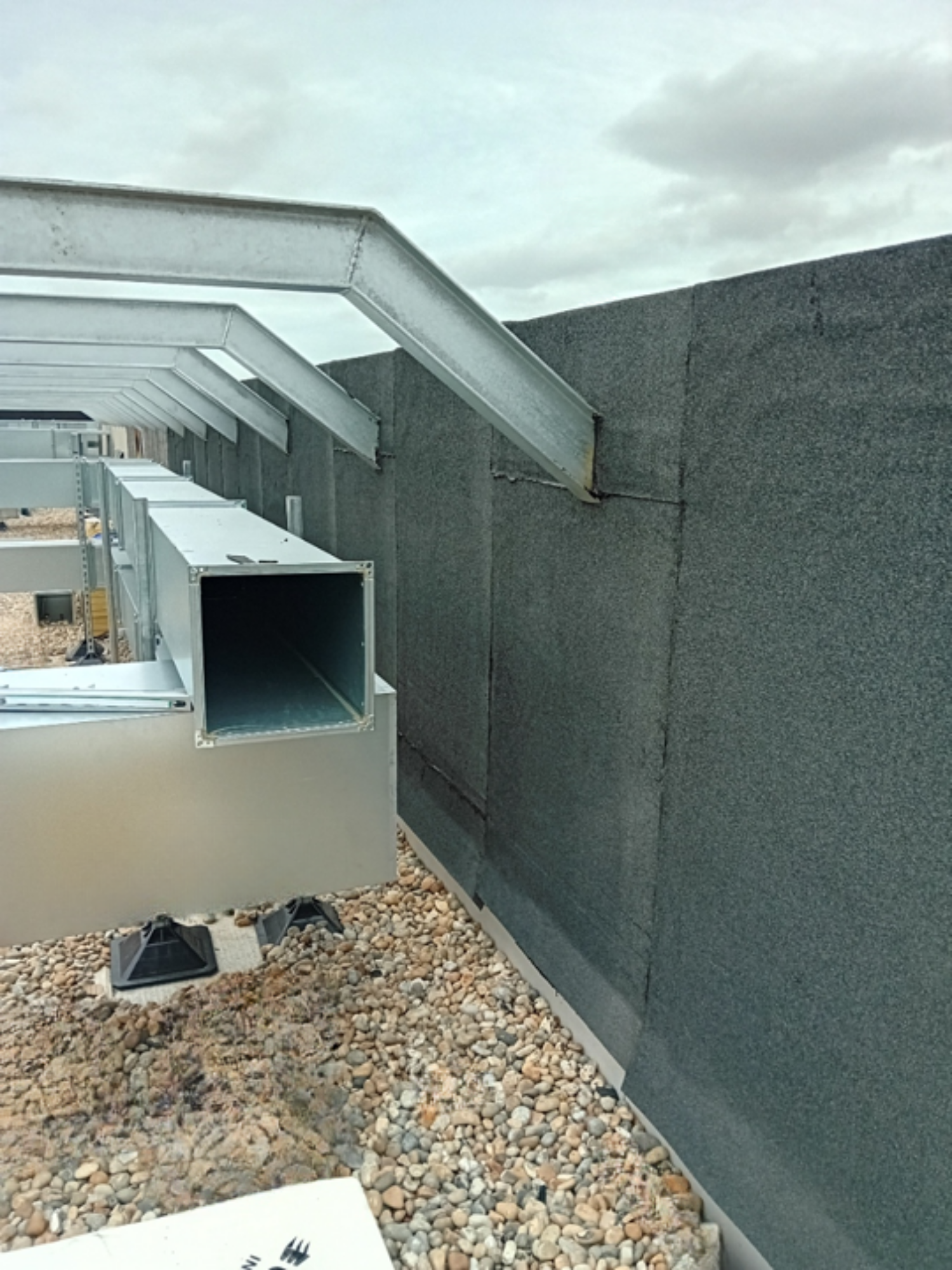 Grey concrete building exterior with white curved roof elements and rectangular opening, gravel ground surface, cloudy sky.