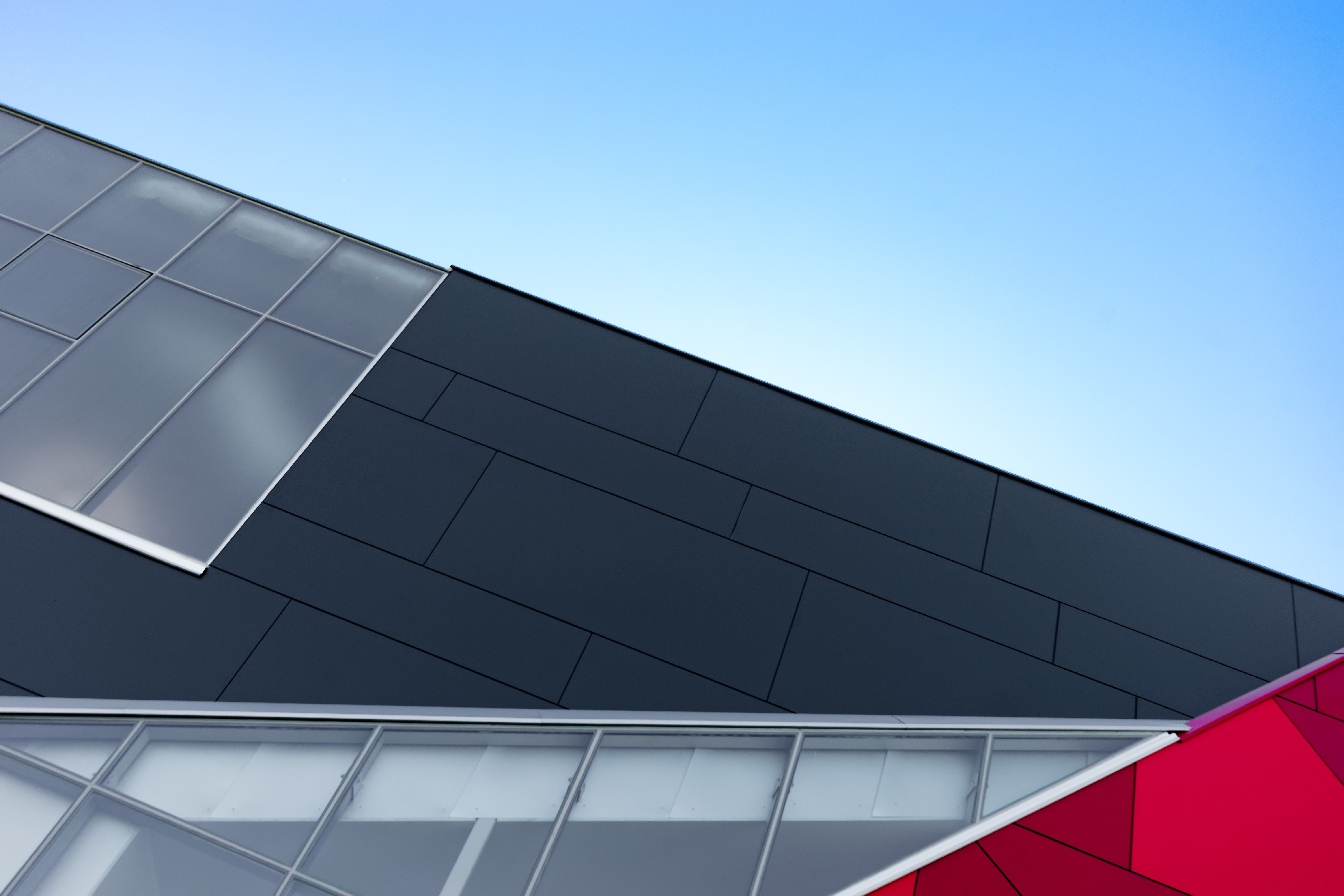 Modern building exterior with angular grey metal panels, red accent section, and glass windows against blue sky.