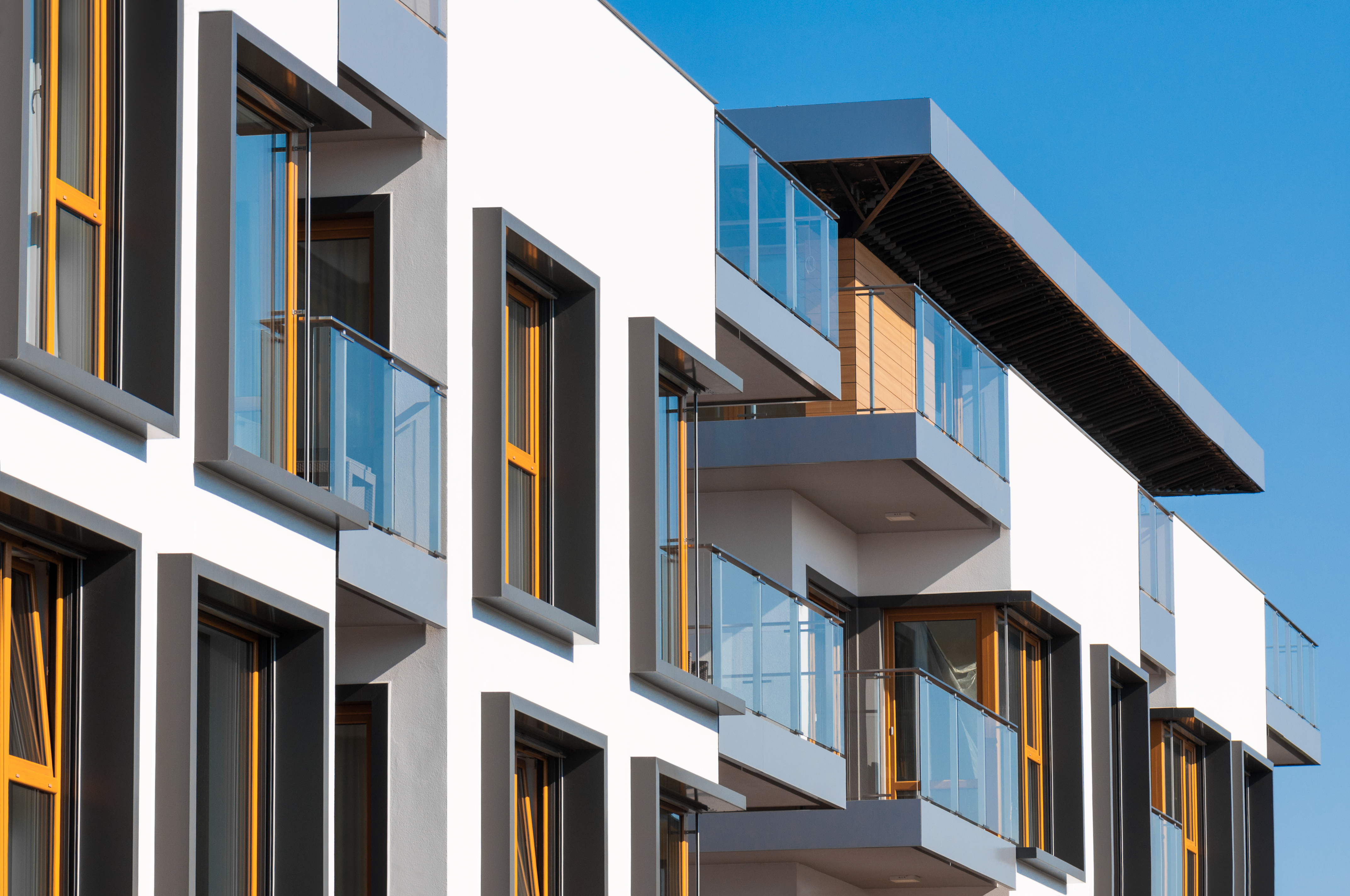 Modern white apartment building with glass balconies, dark window frames, and warm interior lighting against blue sky.