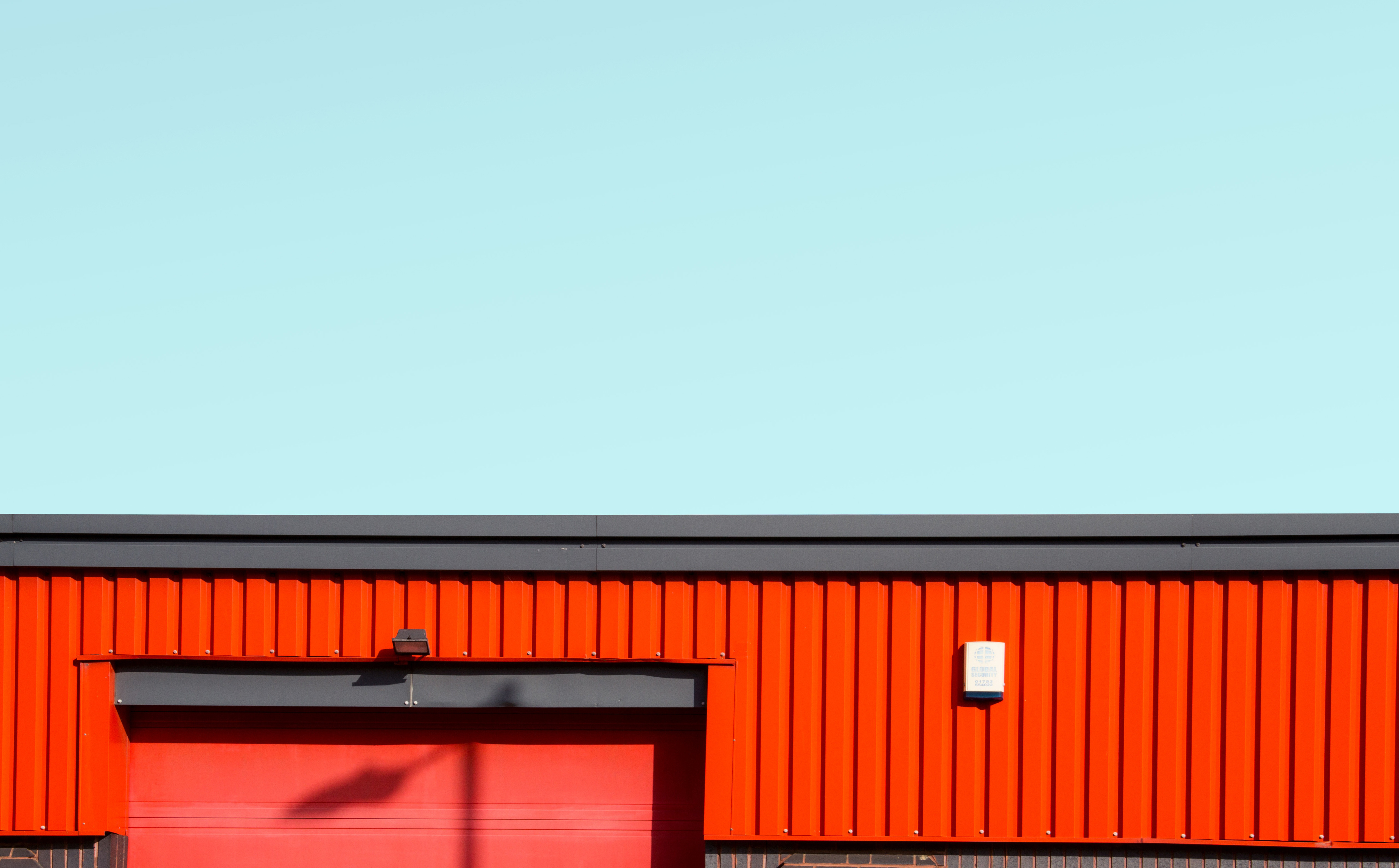 Orange corrugated metal building with grey trim against pale blue-green sky. Red door with dark overhang and small white fixture on wall.