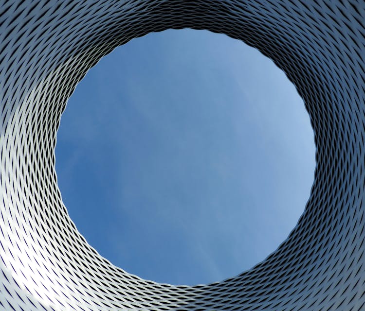 Circular opening framed by radiating metal slats creating concentric pattern, blue sky visible through centre opening.