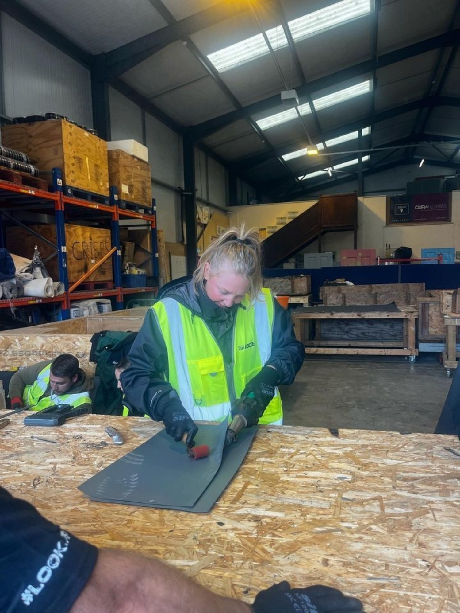 Worker in high-vis jacket cutting dark material on wooden workbench in warehouse with shelving and fluorescent lighting overhead.