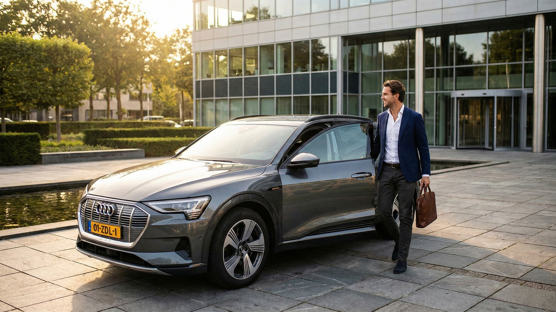 Een glimlachende zakenman in een modern blauw colbert stapt met een aktetas uit een grijze Audi e-tron voor een glazen kantoorpand tijdens het gouden uur.