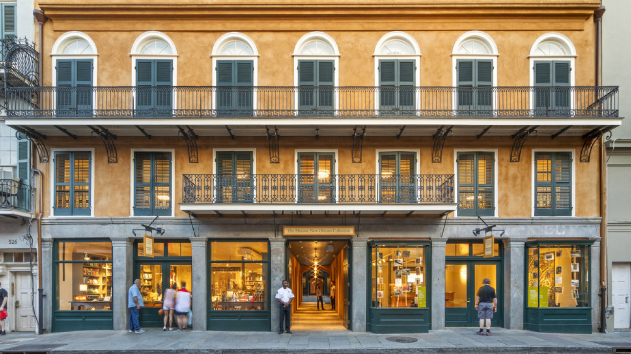 A three-story beige building with green shutters and wrought-iron balconies. The ground floor features large windows displaying various items. Several people stand outside, and an ornate entrance leads inside.
