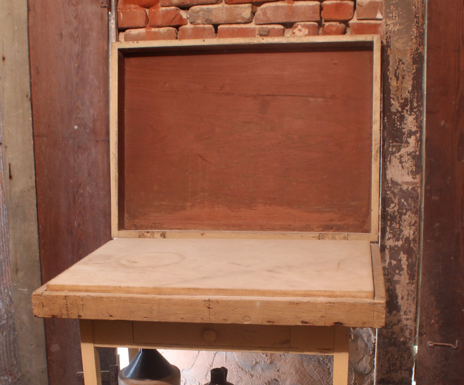 An old wooden desk with an open top, showing a storage compartment. The desk is placed against a brick wall with visible wear and tear, and a rustic, unfinished texture.