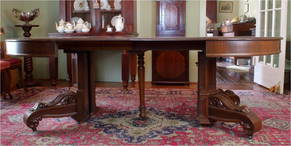 A large wooden dining table with ornate legs sits on a patterned rug in a traditional room. Behind the table, there is a cabinet with porcelain dishes and a wooden door reflecting in a mirror on the floor.