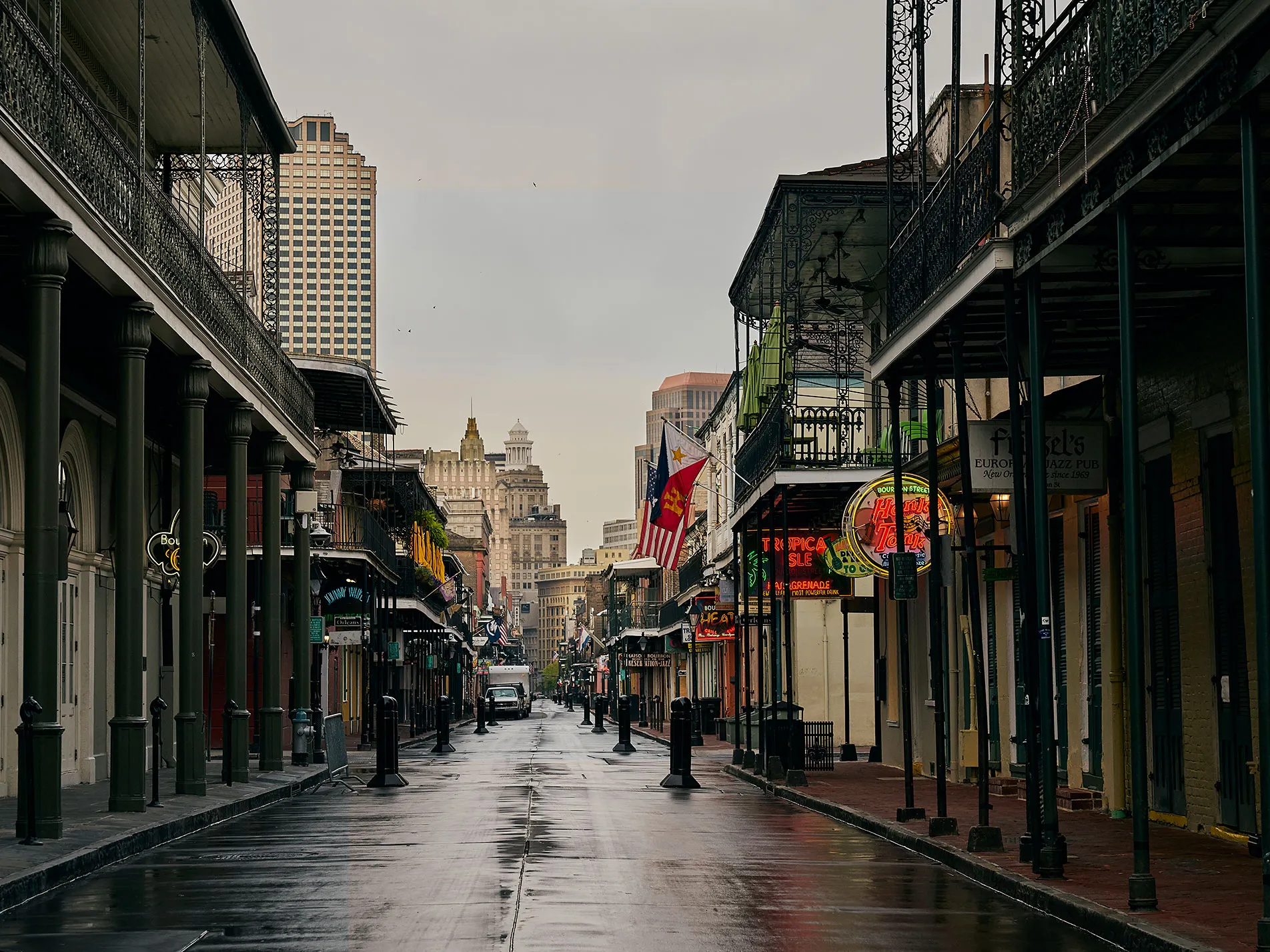 A wet, empty street in New Orleans French Quarter, showcasing old buildings with balconies. Bright neon lights and an American flag add color. Overcast sky and tall skyscrapers are visible in the distance.