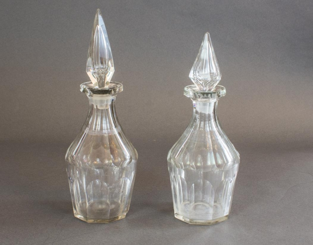 Two glass decanters with pointed glass stoppers, placed side by side against a neutral background. Both have a faceted design and appear empty.