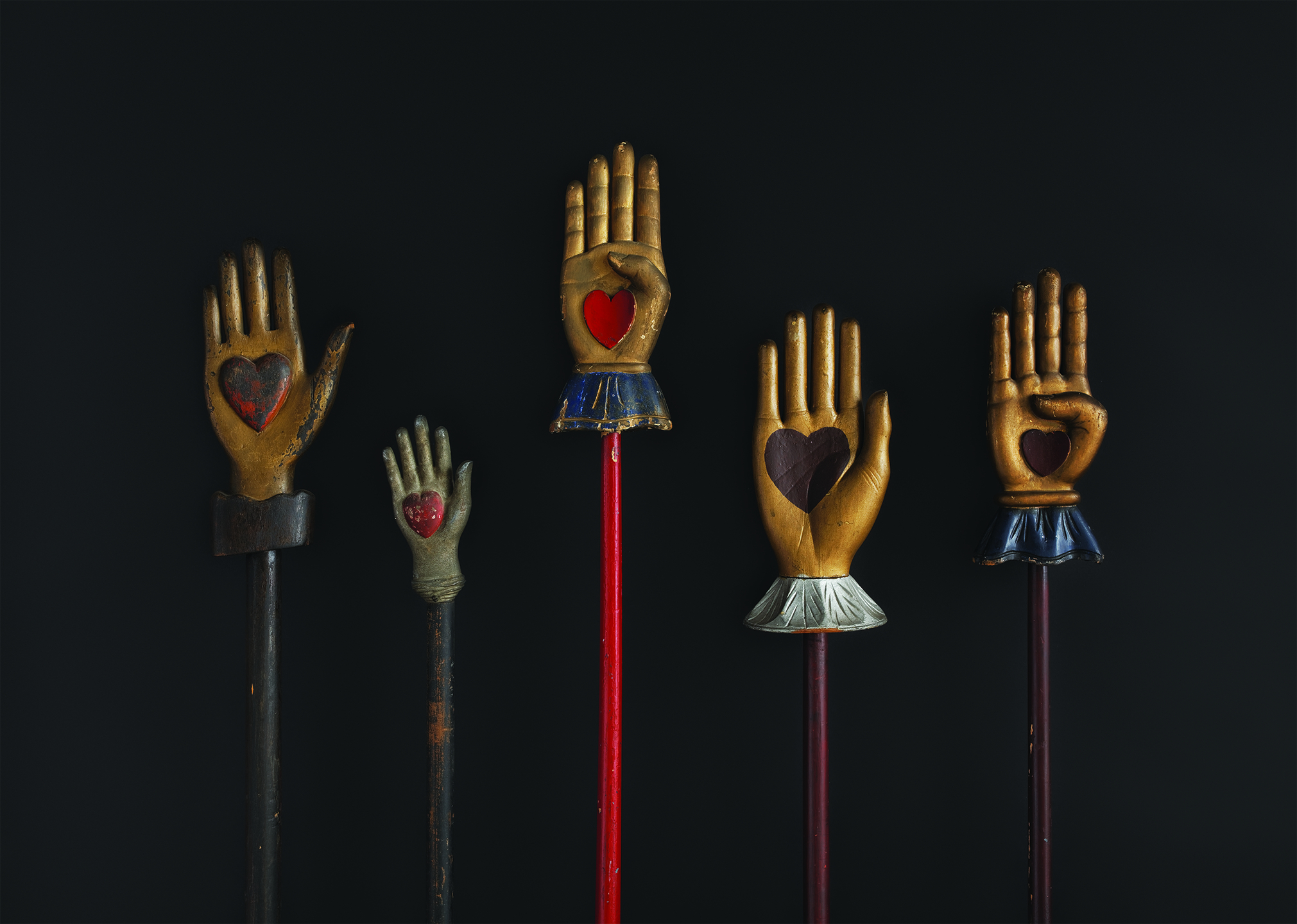 Several decorative hand-shaped staffs against a dark background. Each hand is raised, featuring a heart cutout in the palm and adorned with cuffs in various styles and colors.