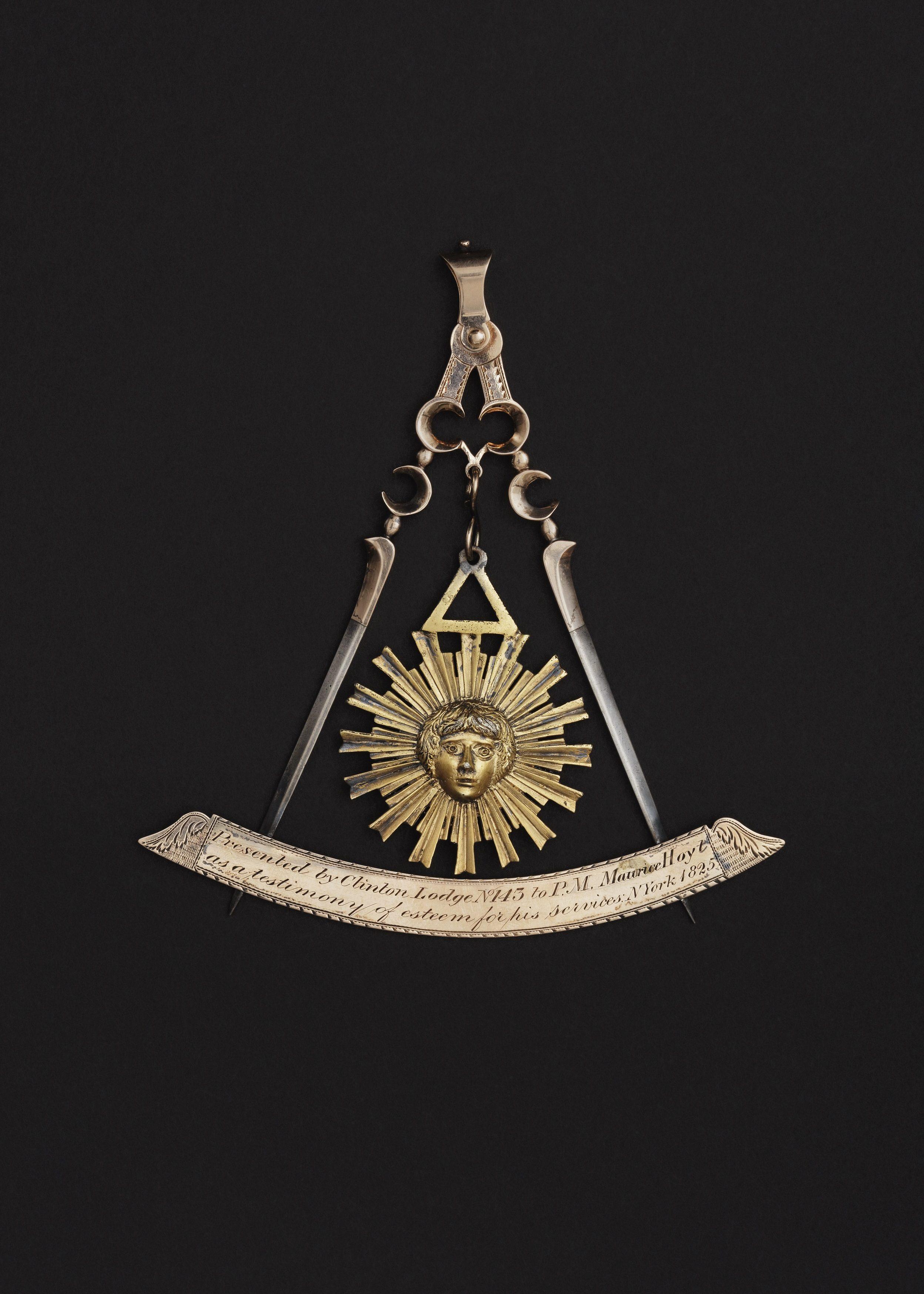 Ornate metallic Masonic emblem featuring a radiant sun face, with a triangular symbol above it. Below, an ornate inscribed scroll is flanked by compasses, set against a black background.
