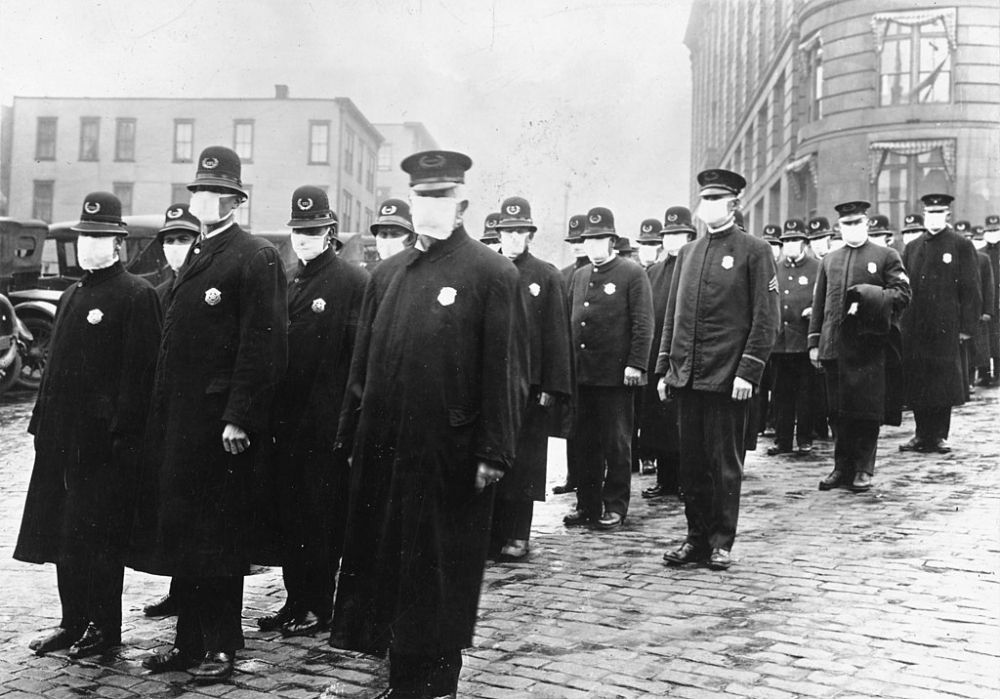 A group of uniformed officers wearing masks stand in rows on a cobblestone street. Buildings and early 20th-century cars are visible in the background, suggesting a historical setting.