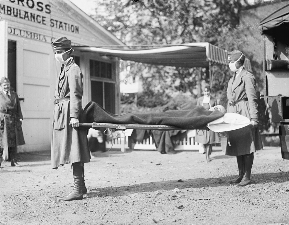 Two women in uniform and masks carry a stretcher with a person covered by a blanket in front of an ambulance station. Other masked individuals are visible in the background, suggesting a historical setting focused on medical response.