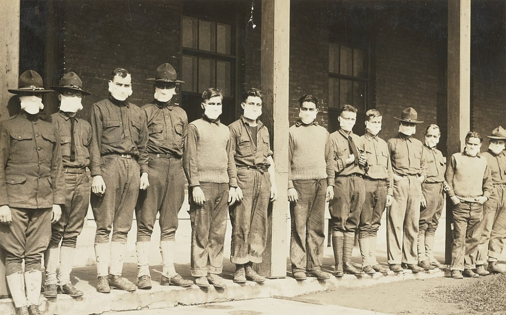 A group of men, dressed in early 20th-century military uniforms and casual clothing, stand in a line on a porch. Many wear cloth masks covering their faces. The background features brick walls and windows.
