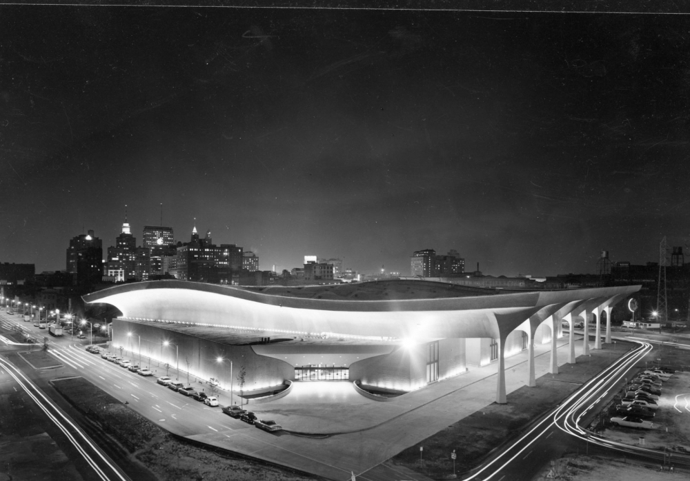 A nighttime aerial view of a futuristic building with a wave-like roof and illuminated columns. City skyline in the background and light trails from passing cars visible around the structure.