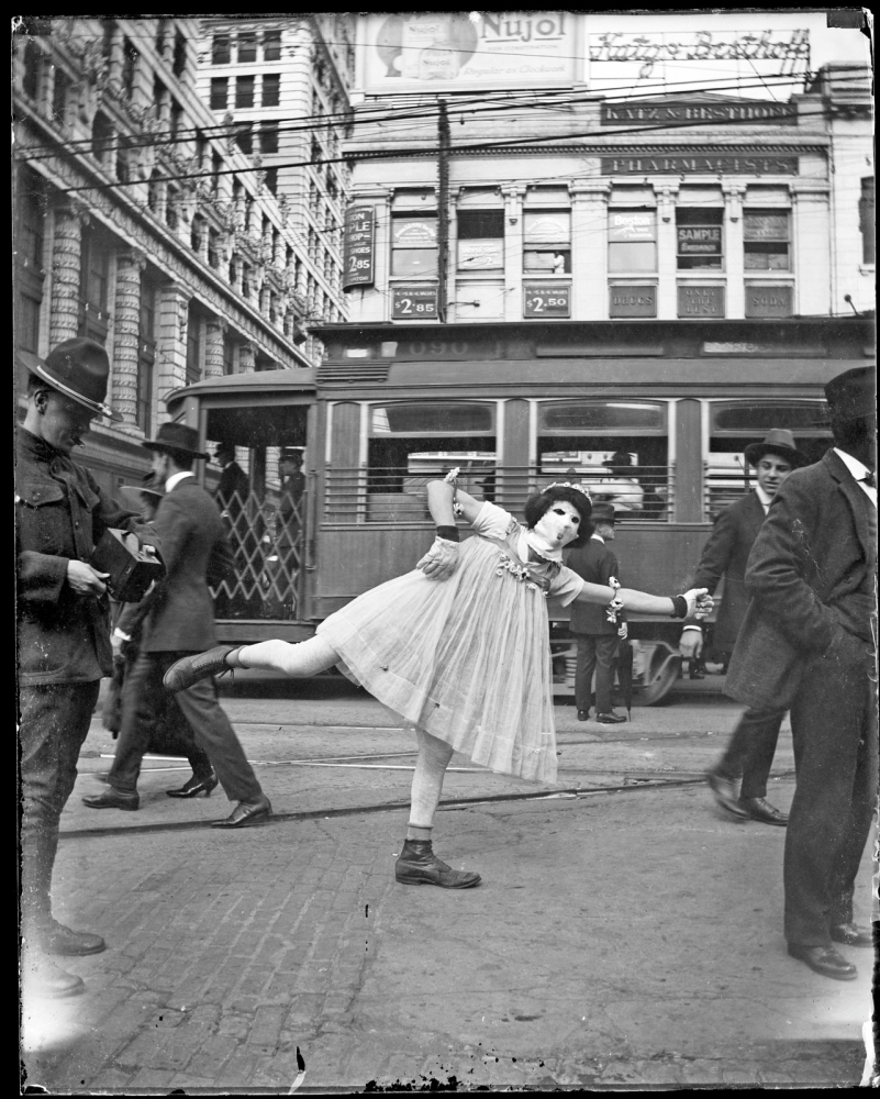 A ballerina poses on one leg in a busy urban street, surrounded by pedestrians and in front of a streetcar. The background shows tall buildings with signs and advertisements. The image is in black and white, capturing a historical moment.