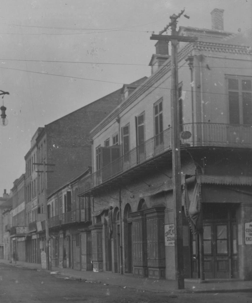 A black and white photo of an old urban street lined with historic buildings. The structures feature balconies and arched windows, characteristic of early 20th-century architecture. The street is empty, and a lamppost stands prominently in the foreground.
