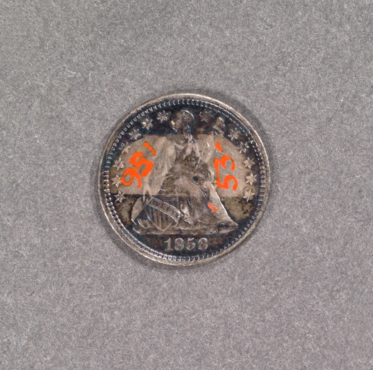 A worn coin from 1858 displaying a seated figure in the center. The coin has visible tarnishing and the numbers 156 and 53 are printed in red over the figure. The background is a plain gray surface.