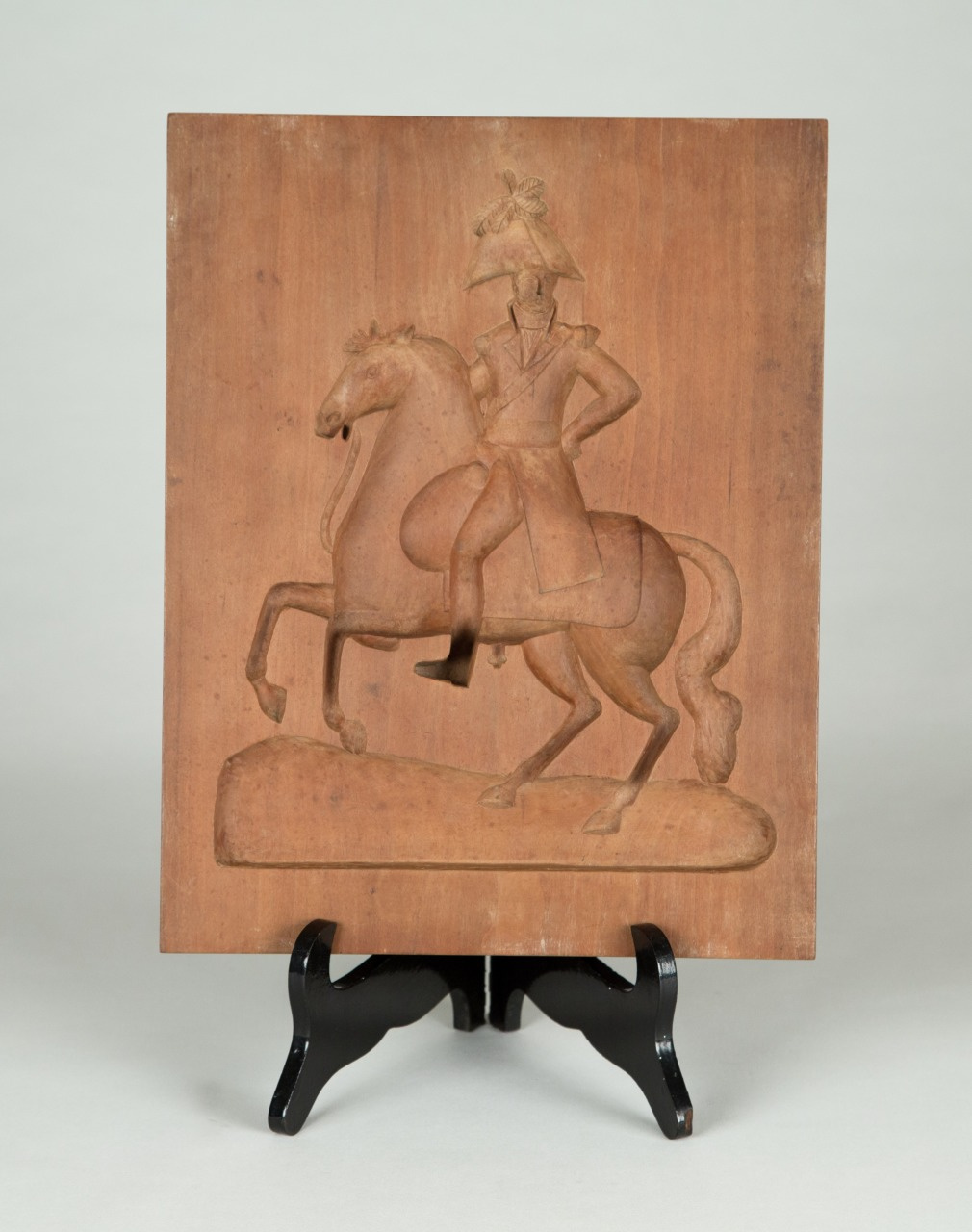 A wooden carving depicts a person in a military uniform riding a horse. The figure is mounted on a rectangular wooden plaque, which is displayed on a black stand.