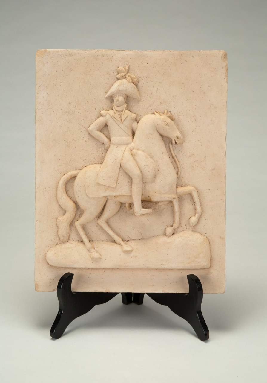 A stone relief sculpture depicting a figure in military attire riding a rearing horse. The relief is mounted on a black stand against a plain background.