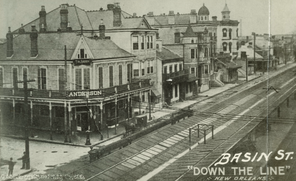 Black and white vintage photograph of Basin Street in New Orleans, featuring a row of historic buildings with streetcar tracks in the foreground. The Anderson building is prominent on the left, and the image is labeled Down the Line.